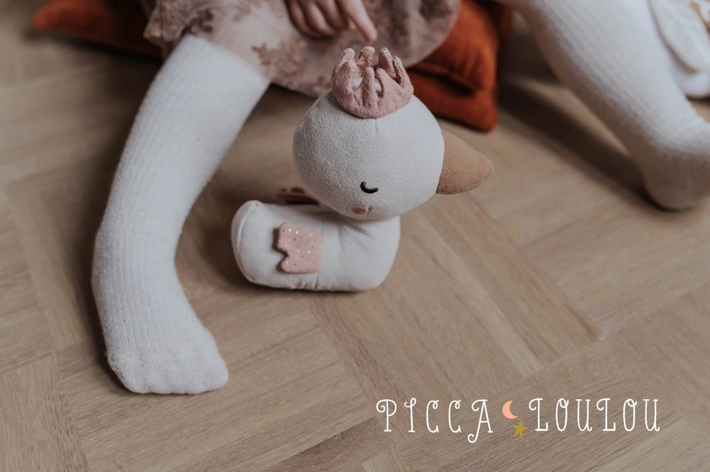 A child in cream-colored tights is sitting on a wooden floor, holding a Duck Davey 17cm plush toy from PICCA LOULOU, adorned with a small pink crown and a pink polka-dotted cape. The floor displays the whimsical text "PICCA LOULOU," making it the perfect nursery decoration.