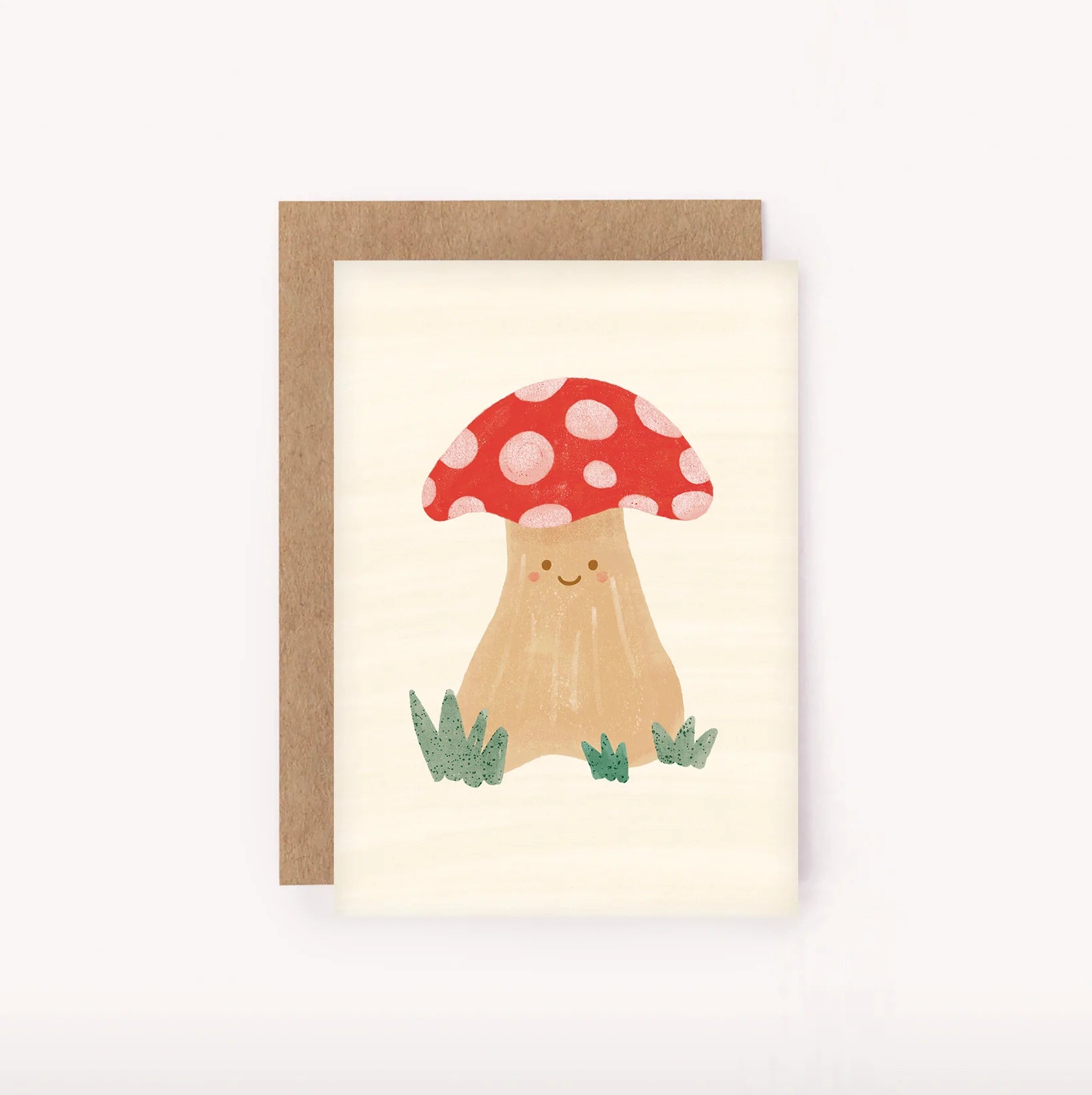 The Mushroom Mini Card by LAUREN SISSONS STUDIO showcases a charming illustration of a smiling mushroom with a red cap and white spots. The mushroom is set against a white background, accented by small tufts of green grass at its base. Made from recycled paper, this delightful mini card comes complete with a brown envelope positioned behind it.