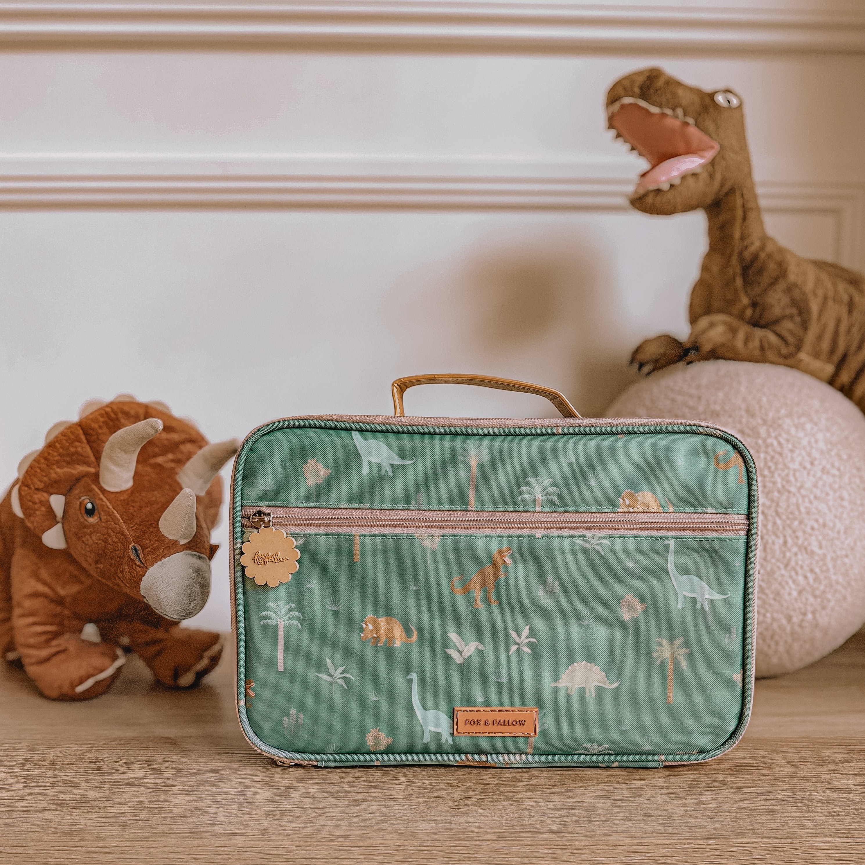 A FOX & FALLOW Dinos Lunch Bag, adorned with a dinosaur pattern and featuring a brown leather handle and a small charm hanging from the zipper, sits on a wooden surface. Behind it, there are two plush dinosaur toys. A white wall with molding is in the background.