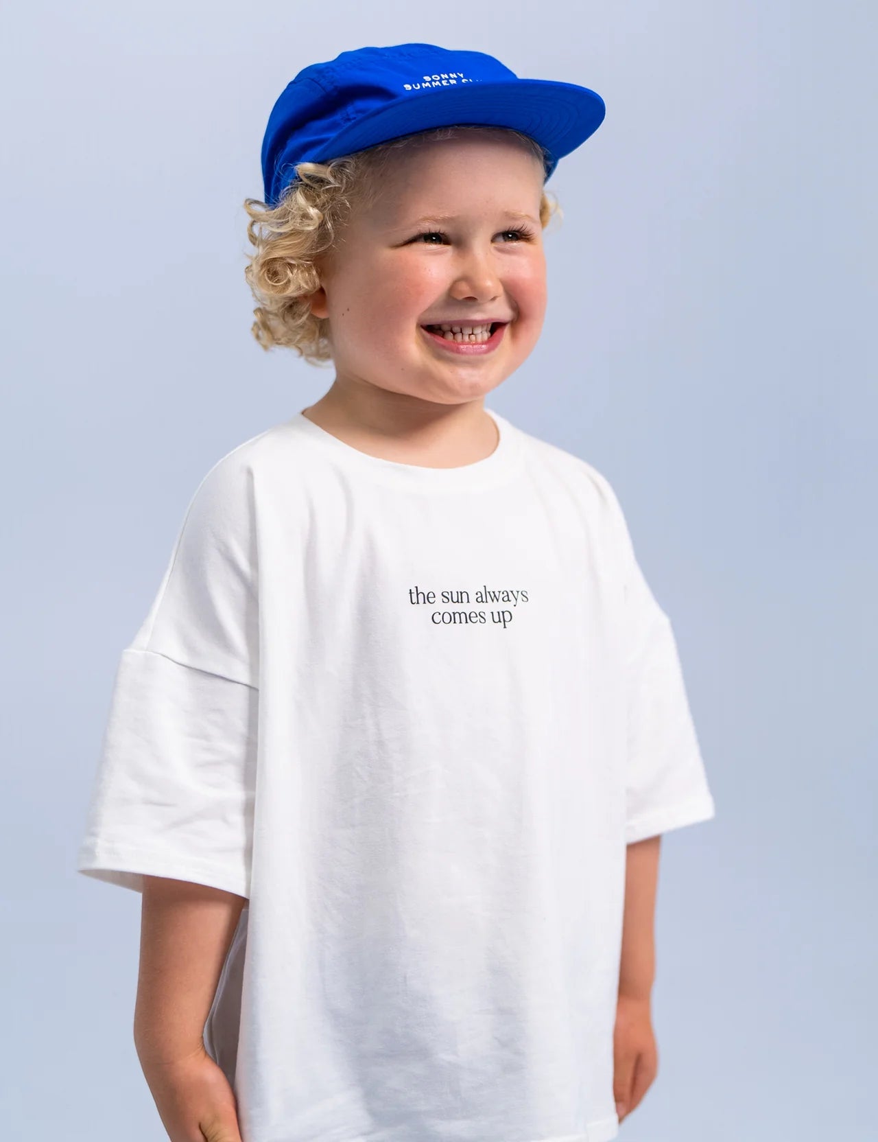 A smiling child with curly hair is wearing the SONNY LABEL Summer Club Cap : Electric, which features an adjustable velcro strap, and a white T-shirt that says "the sun always comes up." The background is solid light blue.