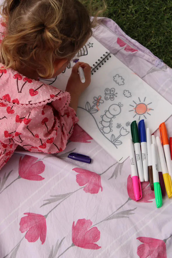 A child in a pink dress lies on a blanket outside, exploring their creativity with the OH MY FRANKIE Oh My Scribbles Reusable Colouring Book. The drawing features a caterpillar, sun, butterfly, and flowers, with markers scattered around to spark imagination.