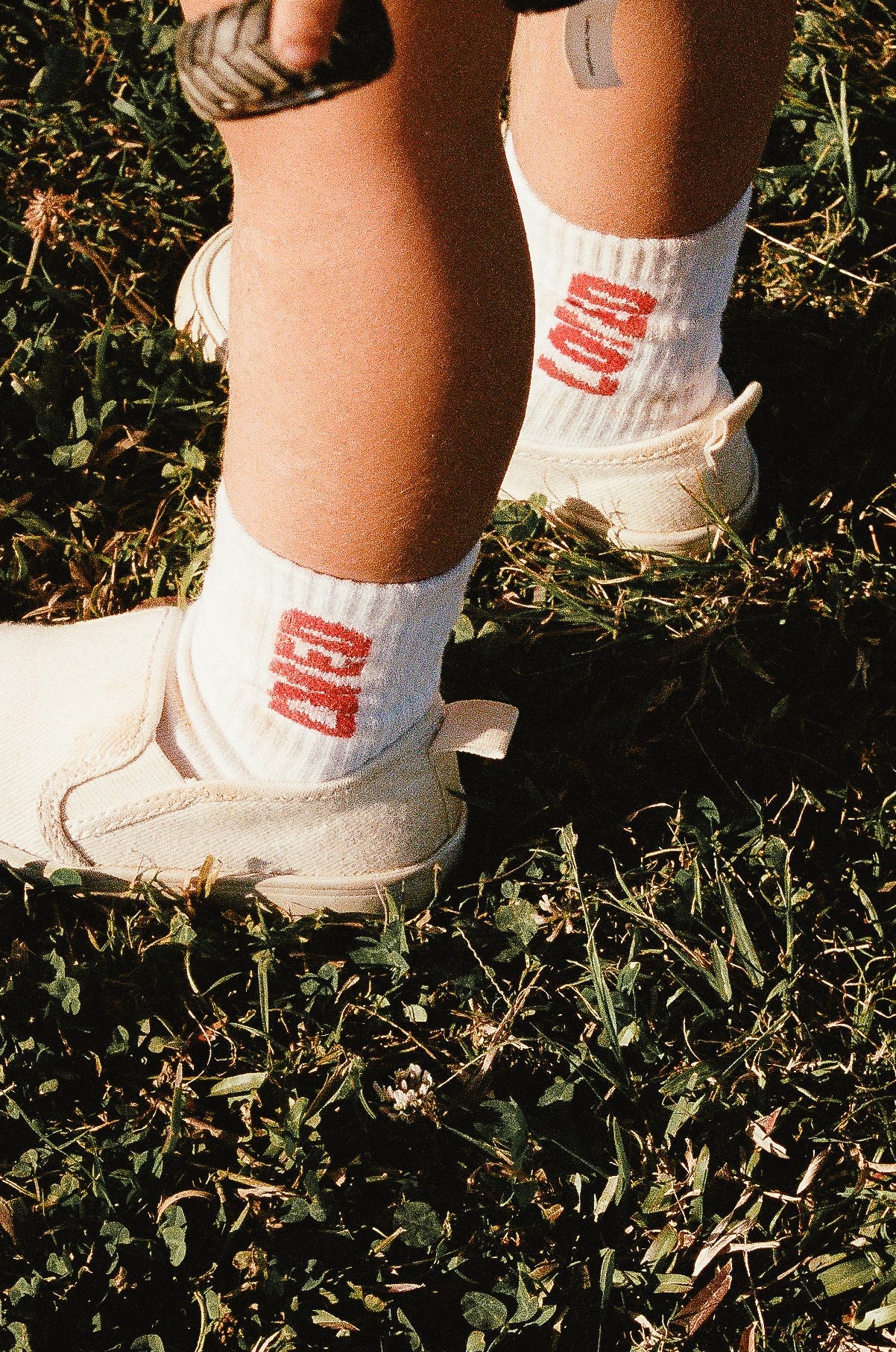 Close-up of a 2-5 year old child's legs standing on grass. The child is wearing TINY LOVE CLUB's Loved Socks, featuring white sports socks with red letters and elastic banded ankles, paired with white shoes. They are holding an object in their right hand.