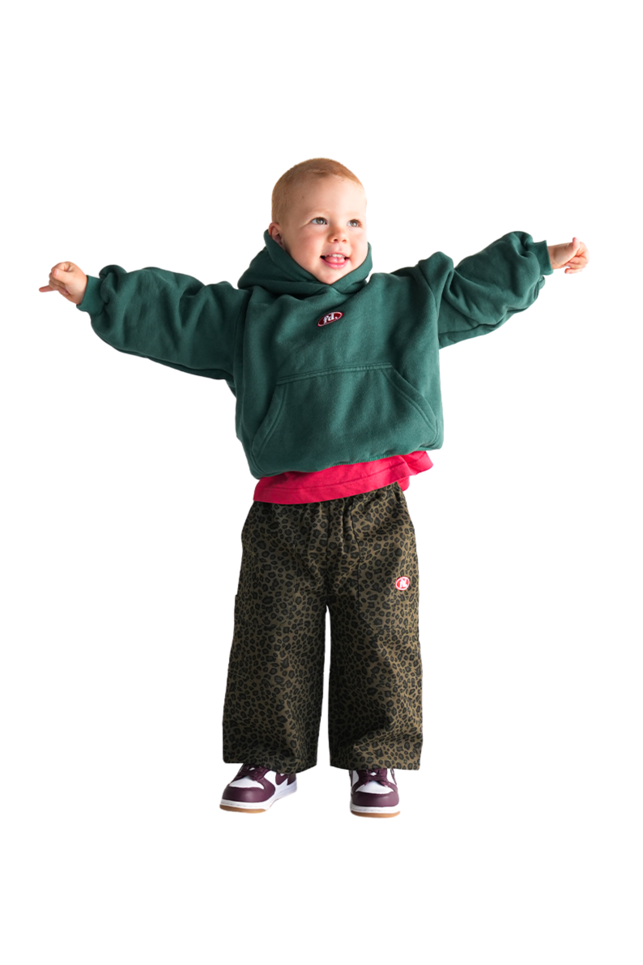 A joyful child smiles against a white background wearing a green hoodie, purple sneakers, and a red shirt beneath. They're standing with arms outstretched in FRANCO'S DAD's PRE-ORDER Leopard Cargos.