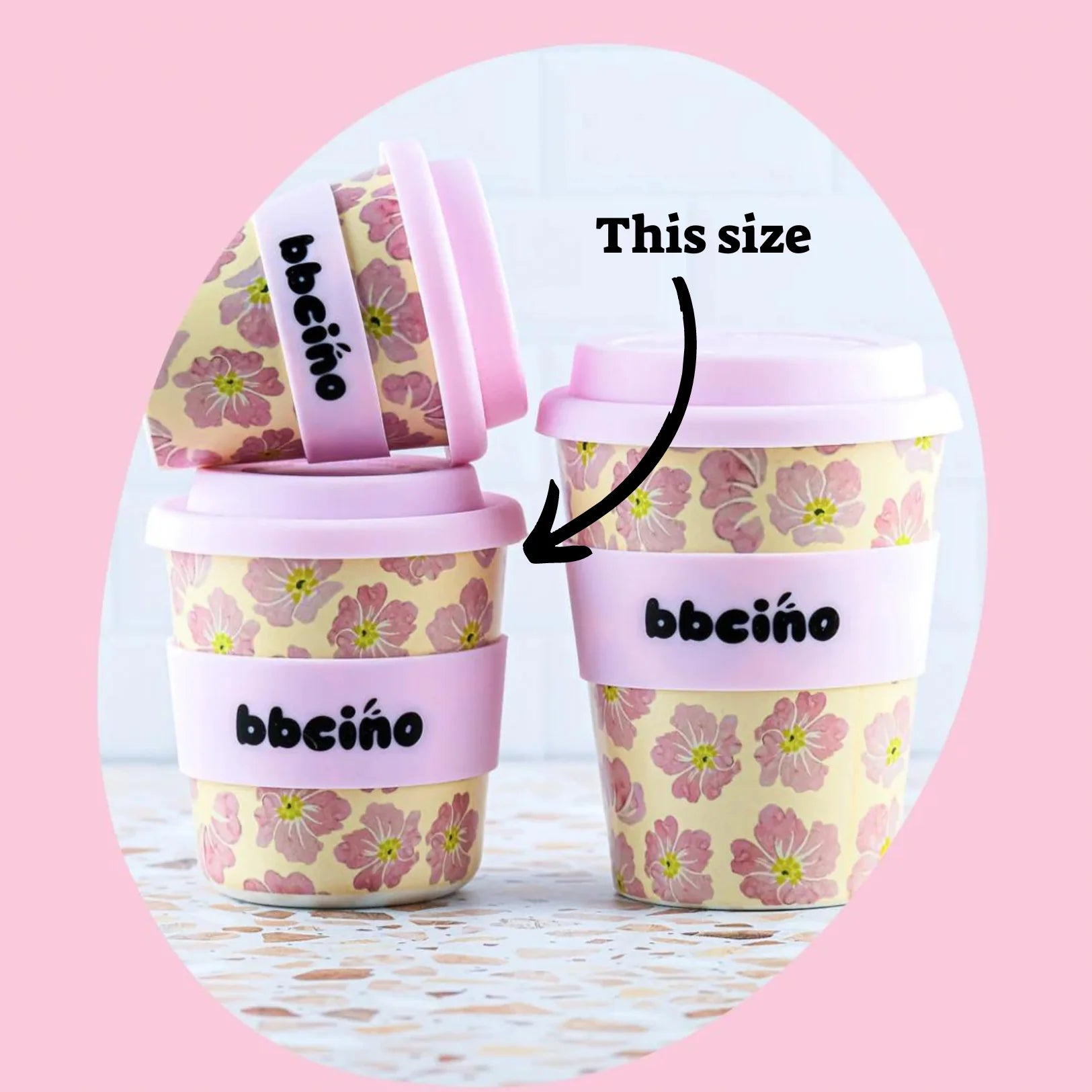 Three BBCINO reusable coffee cups in pink with a floral pattern are arranged against a light pink background. The two smaller cups are labeled with an arrow pointing to one cup, indicating "This size" in black text. The midi-sized cups, named Reusable BambinoCino Cup 240ml Poppy, are branded with "BambinoCino" on the lids and sleeves.