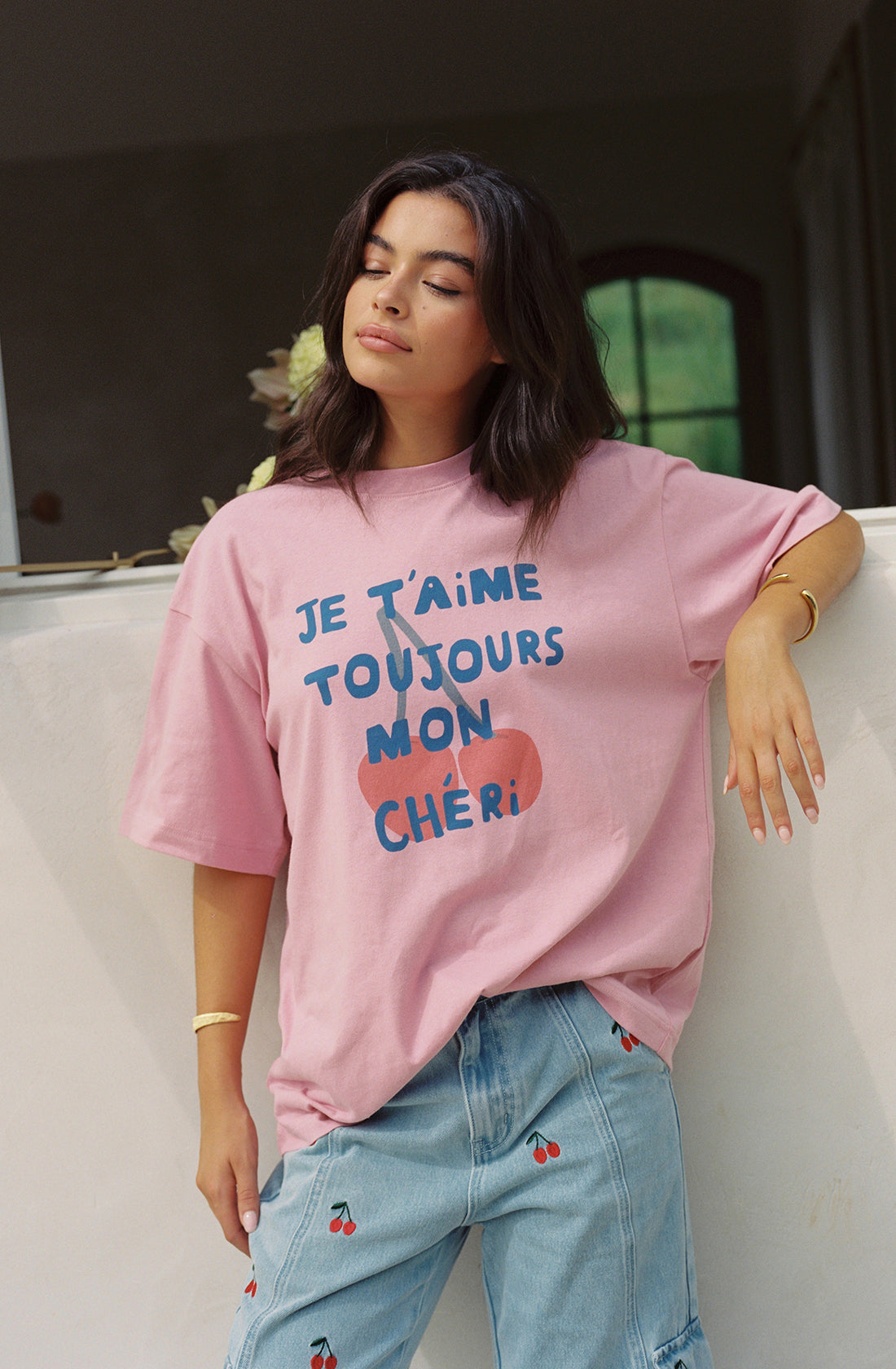 A person with long dark hair stands with eyes closed, leaning against a white ledge. They are wearing an oversized fit, Je T’aime Tee from THE WHOLESOME STORE featuring blue text that reads "JE T'AIME TOUJOURS MON CHÉRI," paired with light-wash jeans adorned with small cherry prints. The outfit is GOTS certified and ethically made.