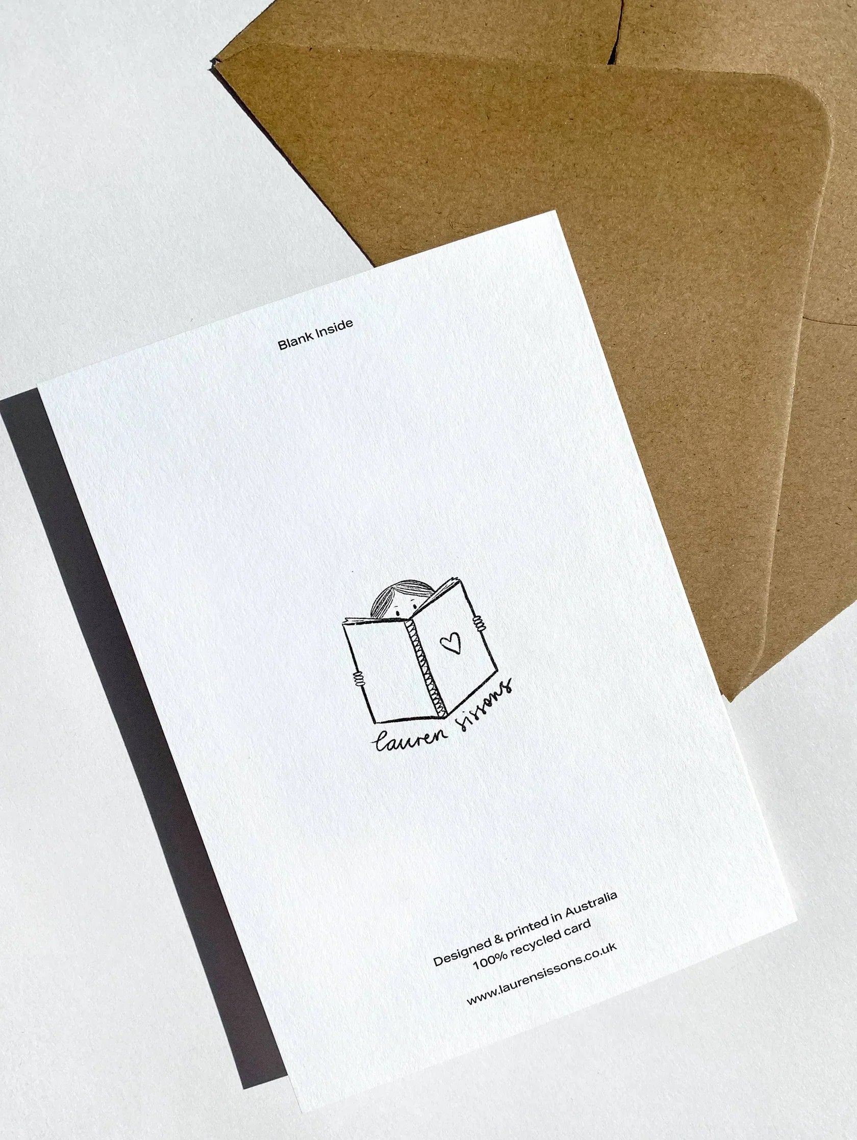 A greeting card against a brown envelope background. The Mushroom Mini Card, printed on recycled paper, features a simple illustration of a person holding an open book with a heart on the cover and the text "LAUREN SISSONS STUDIO" below. The card text reads: "Blank inside," "Design by Printed in Australia," and includes a web address.