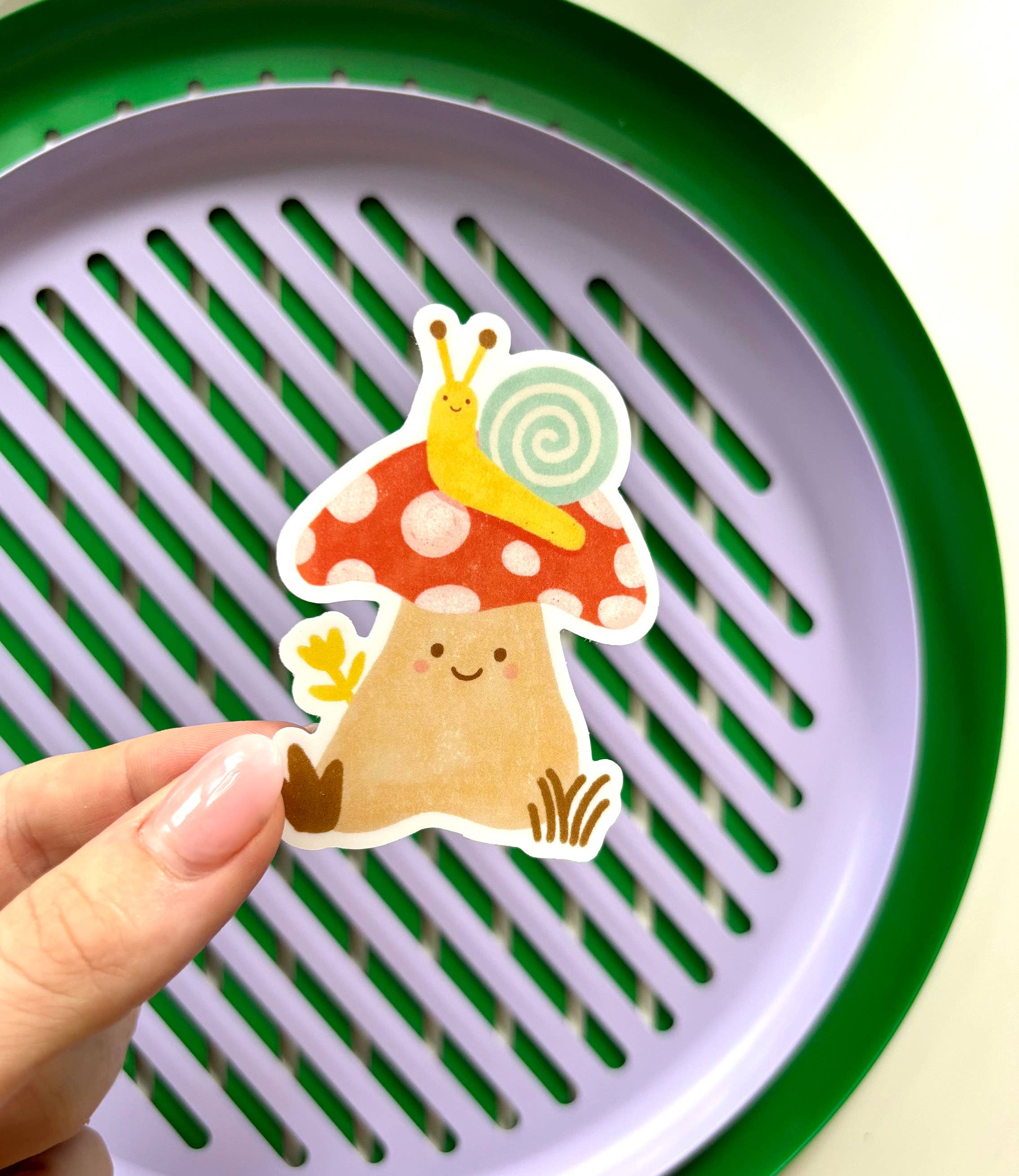 A hand holds the Mushroom Sticker from LAUREN SISSONS STUDIO, showcasing a cute tan mushroom with red spots, a smiling face, and a small snail with a yellow shell on top. The background features a green and purple circular grater.