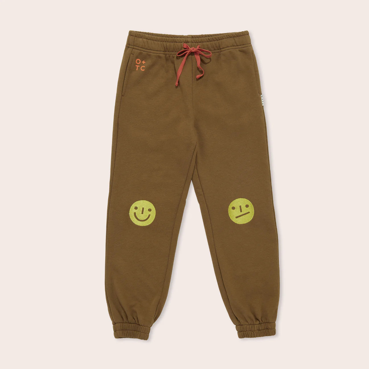 The Mood Track Pants by OLIVE + THE CAPTAIN are brown sweatpants made from premium cotton fleece. They feature a red drawstring and elastic cuffs, along with fun mood faces—embroidered smiley faces on the knees, one happy and one neutral. Additionally, there is small text on the left hip that reads "O + T C.