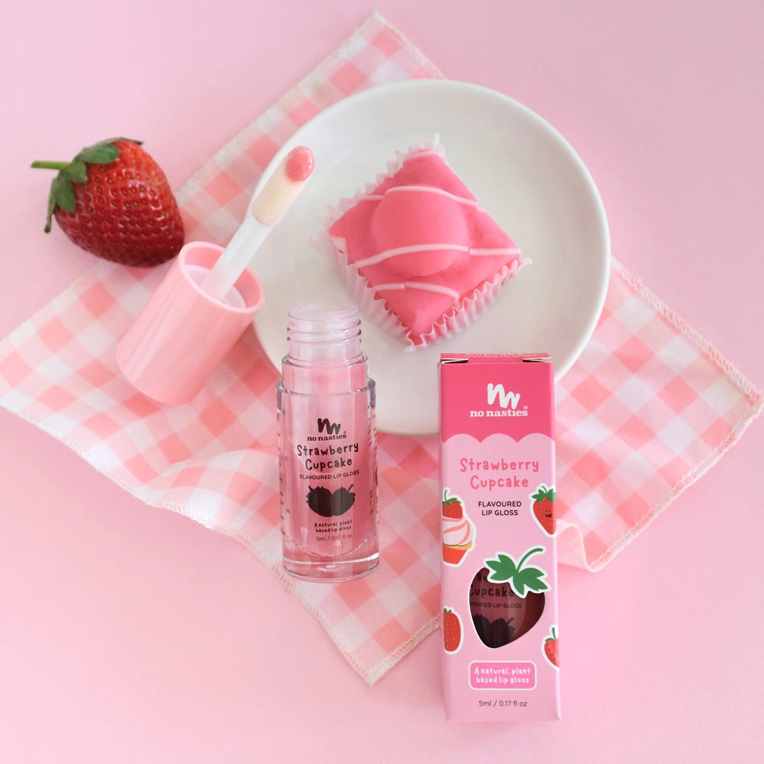 The NO NASTIES Natural Kids Lip Gloss Wand in Strawberry Cupcake/Pink, featuring strawberry and cupcake illustrations on the box, is displayed with a pink cupcake and a strawberry on a white plate atop a pink gingham napkin.