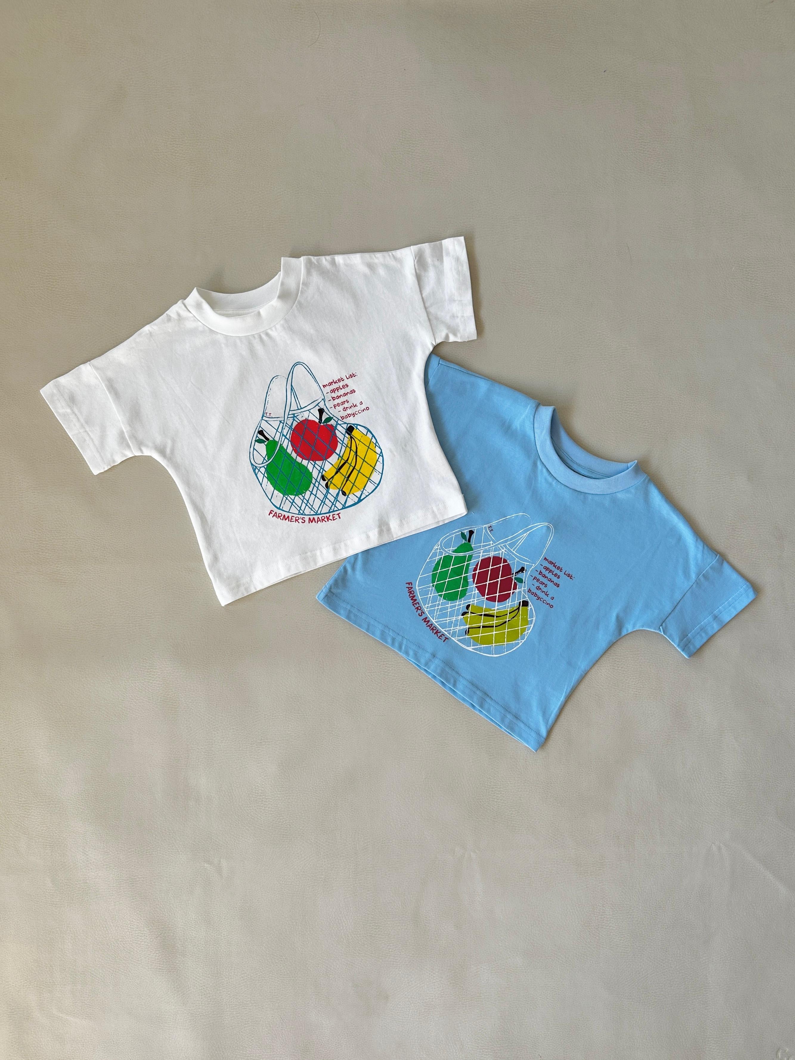 Two children's T-shirts from TINY TROVE are laid flat on a neutral background. One is the Farmer's Market Relaxed Tee in white, and the other is blue. Both feature a colorful fruit illustration with the text "AVOCADO MARKET.