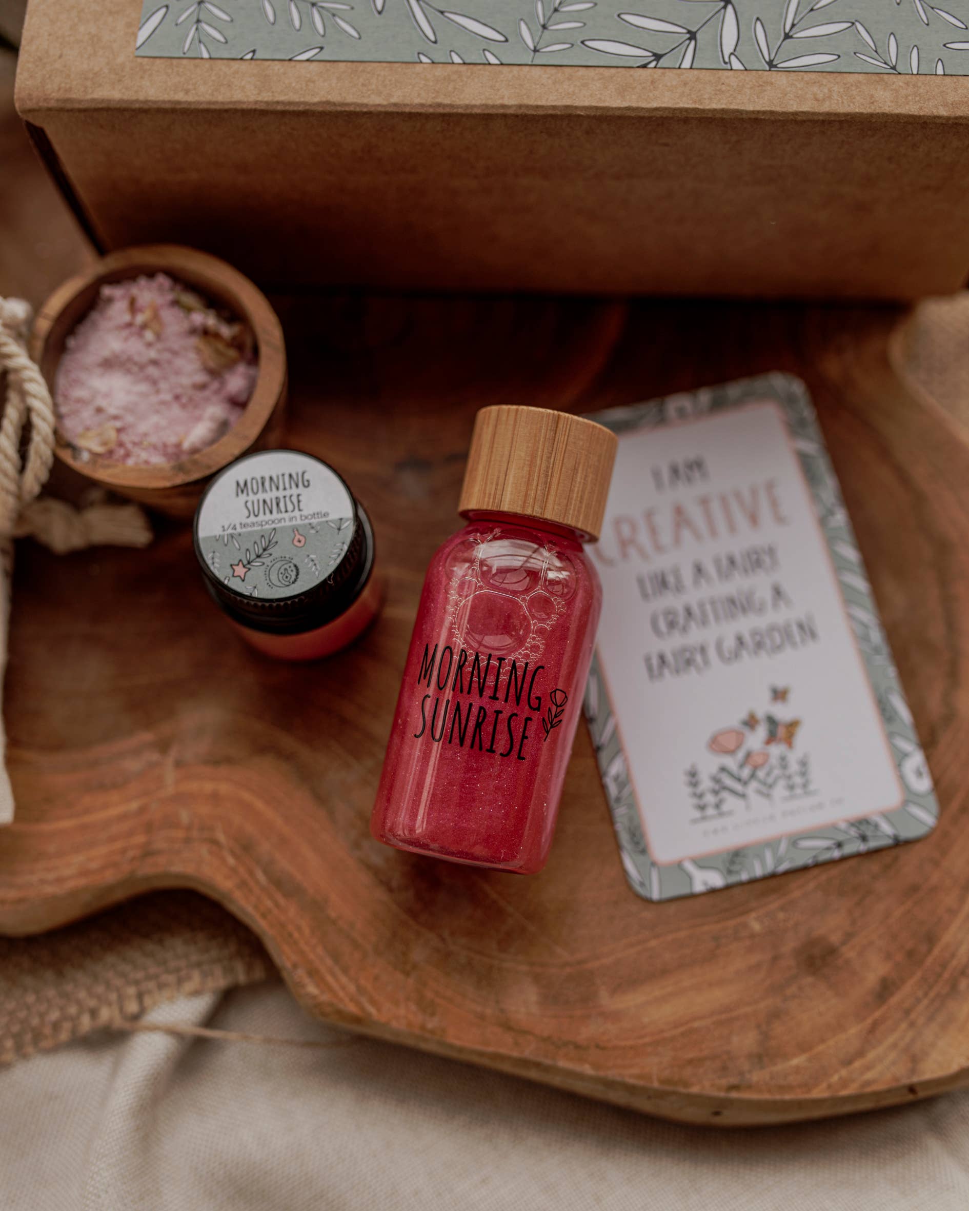 Flat lay of a wooden surface featuring a small jar of red liquid labeled "Morning Sunrise," a tub of pink scrubbing crystals, and a decorative-edged card with an inspirational quote about creativity next to the patterned box lid from THE LITTLE POTION CO's MINI Enchanted Garden Potion Kit (A potion for creativity).