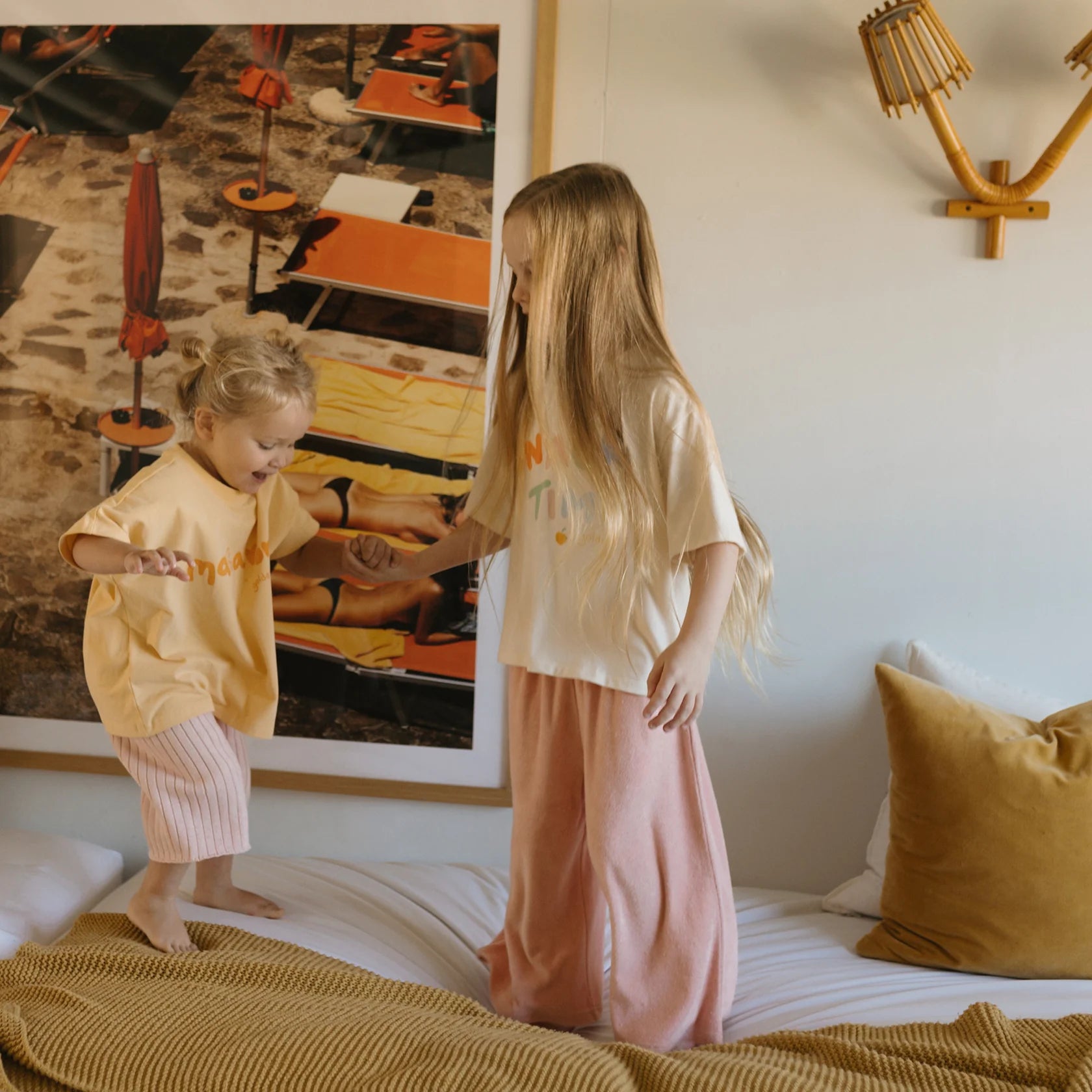 Two children are standing on a bed in a room with beige and brown decor. The older child, with long blonde hair, sports a white shirt paired with Confetti Wide Leg Pants Pink Parfait from GOLDEN CHILDREN. The younger child, donning a yellow shirt and pink shorts, stands beside them. An artwork and light fixture adorn the wall behind them.