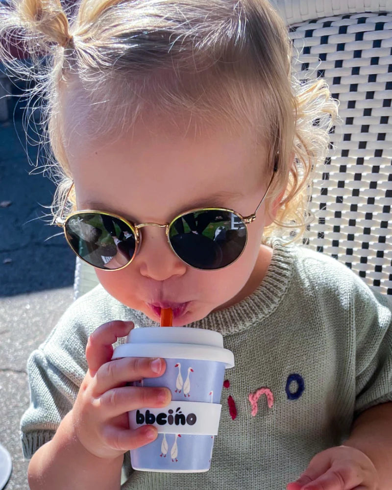 A young child with curly hair and sunglasses sips from a small reusable cup adorned with playful geese and the "BBCINO" brand. Crafted from recyclable silicone, this sustainable Babycino cup, known as the 120ml Loosey-Goosey, perfectly complements the child's light green sweater adorned with colorful embroidery.