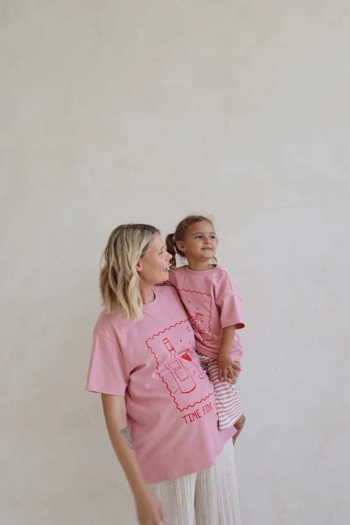 A woman cradles a child as they wear matching oversized pink tees from BOBBY G BABY WEAR's "Time For A Wine" collection. The child, in striped pants and looking elsewhere, adds to the cozy vibe against a plain, light-hued wall.