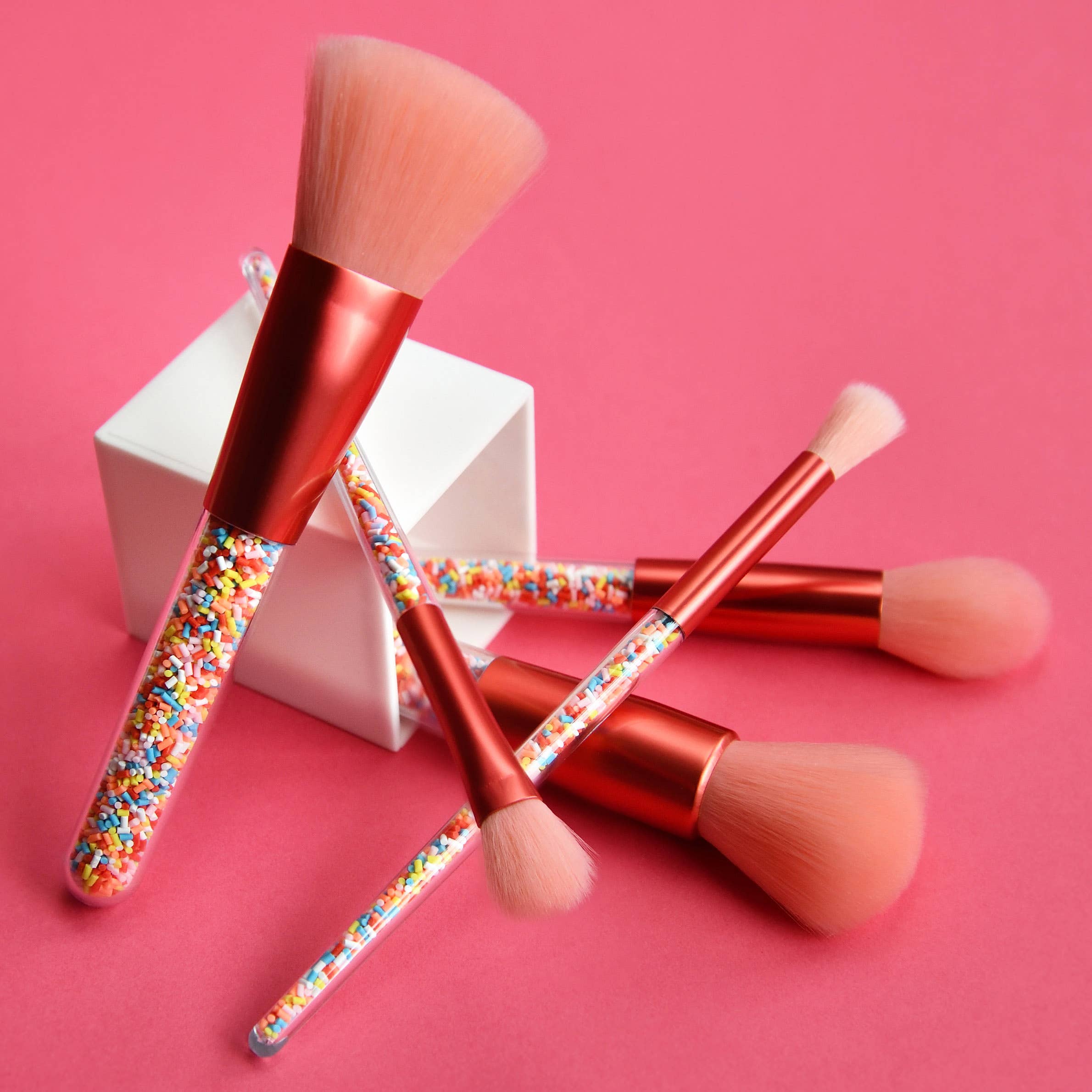 The Twinkle Sprinkle Brush Set by no nasties kids, a vegan and cruelty-free makeup brush set featuring pink bristles and red handles filled with colorful sprinkles, is artfully arranged on a pink background. The hypoallergenic brushes are displayed artistically, with one resting against a small white cube.