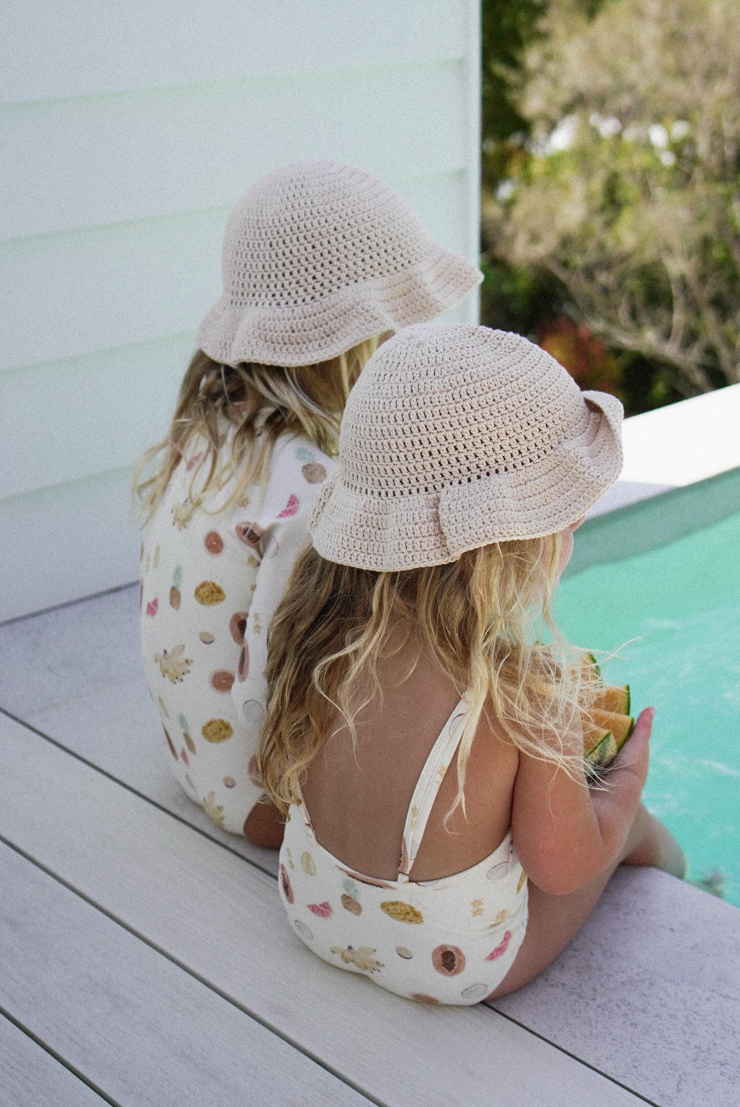 Two young children sit on a wooden deck next to a pool, facing away from the camera. Both wear matching, cream-colored crochet sun hats and light-colored Tutti Frutti One Pieces by EN. THE LABEL, adorned with playful patterns. The background includes a white wall and blurred greenery.