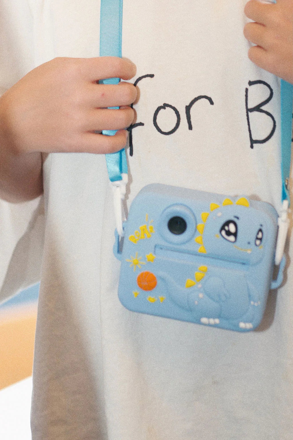 A person wearing a light-colored shirt with "for B" partially visible holds the Oh My Frankie ~ Oh My Snap Instant Print Camera Blue, featuring a vibrant yellow and orange cartoon dinosaur design. Perfect for creative play, it dangles from a matching blue wrist strap.