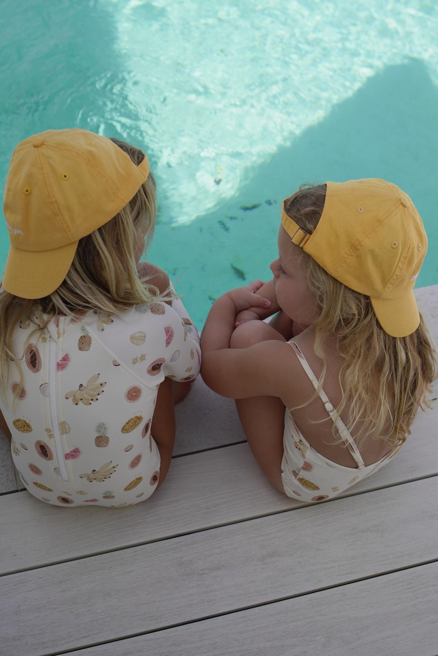 Two young children with long blonde hair sit side by side by a clear blue pool. They both wear yellow baseball caps and white toddler swimsuits with colorful patterns. One child wears the "Tutti Frutti One Piece" swimsuit from EN. THE LABEL that features sleeves, while the other sports a sleeveless variation of the same brand's one-piece, featuring adjustable straps.