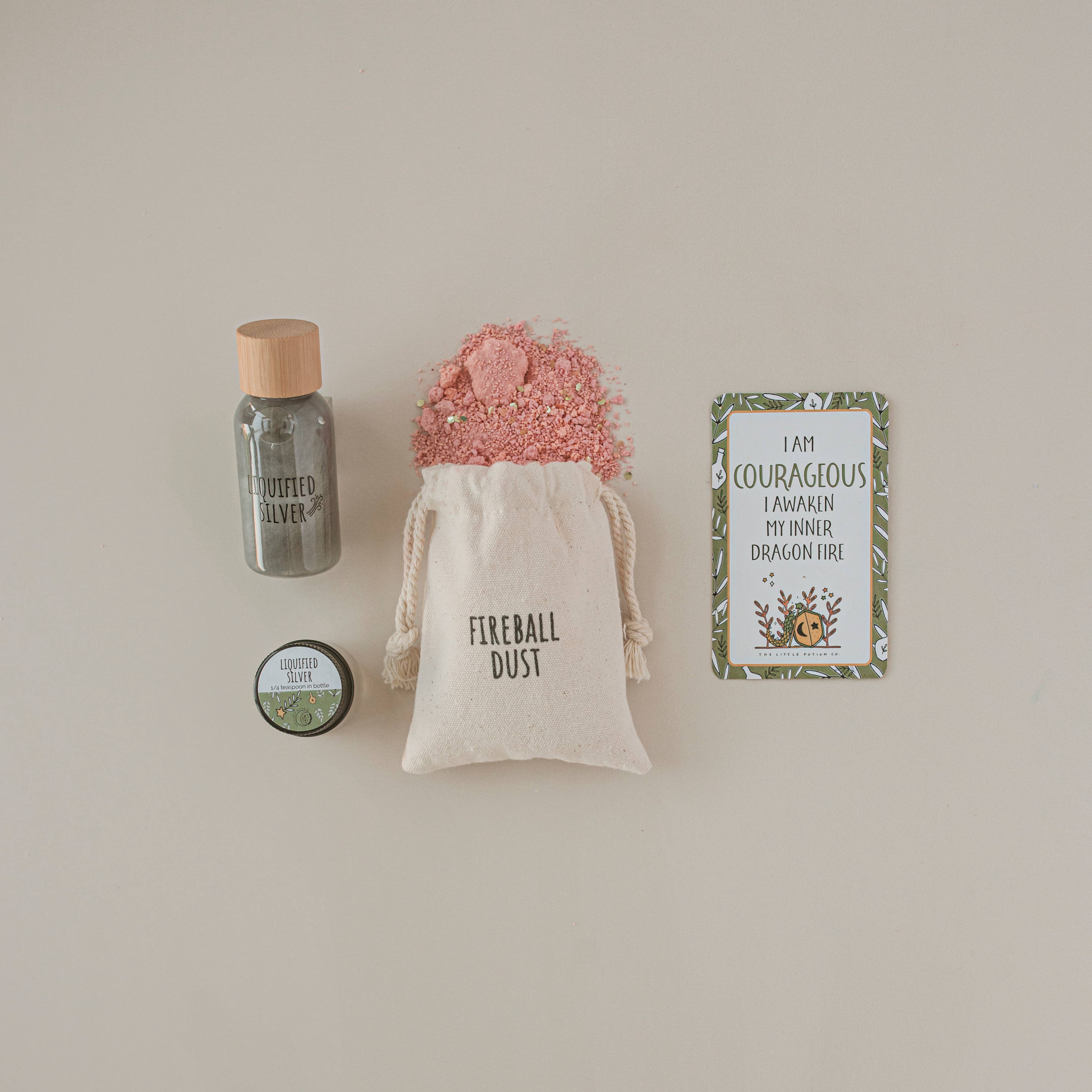 A small bottle of glitter labeled "Silver," a tin marked "Enchanted Elixir," and a cloth bag with "Fireball Dust" spilling out are arranged alongside a card reading, "I am courageous, I awaken my inner dragon fire," creating the perfect MINI Dragon Tales Potion Kit by THE LITTLE POTION CO for imaginative play on a beige background.