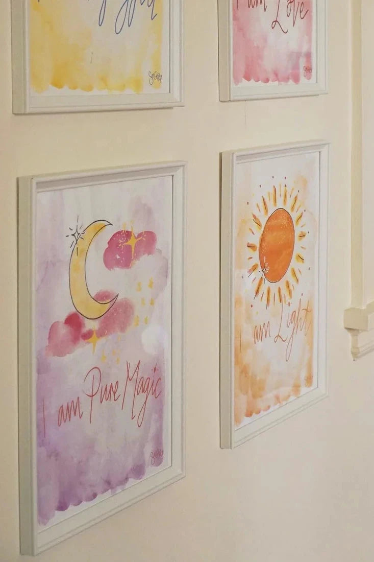 A cozy room corner showcases an A4 "I am Pure Magic" Art Print from SUN CHILD, featuring hand-painted watercolor illustrations. Phrases such as "I am Pure Magic" and "I am Light" grace the walls, while plush toys, including a pink hippo, sit happily on the windowsill.