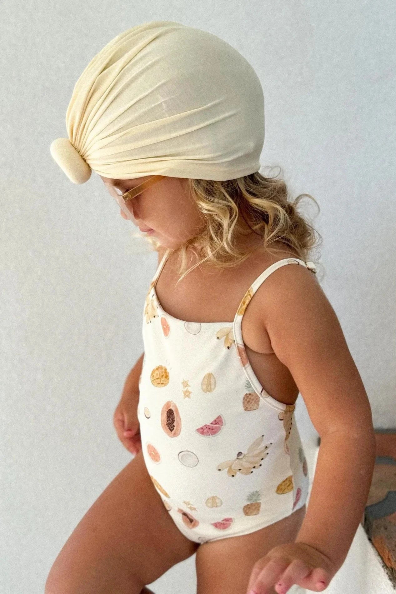 A young child with blonde hair wears a light yellow turban headwrap, sunglasses, and the Tutti Frutti One Piece by EN. THE LABEL, which is a white toddler swimsuit adorned with various pastel-colored shapes and animal prints. The child looks downward and appears to be standing against a plain backdrop, showcasing the adjustable straps for a perfect fit.