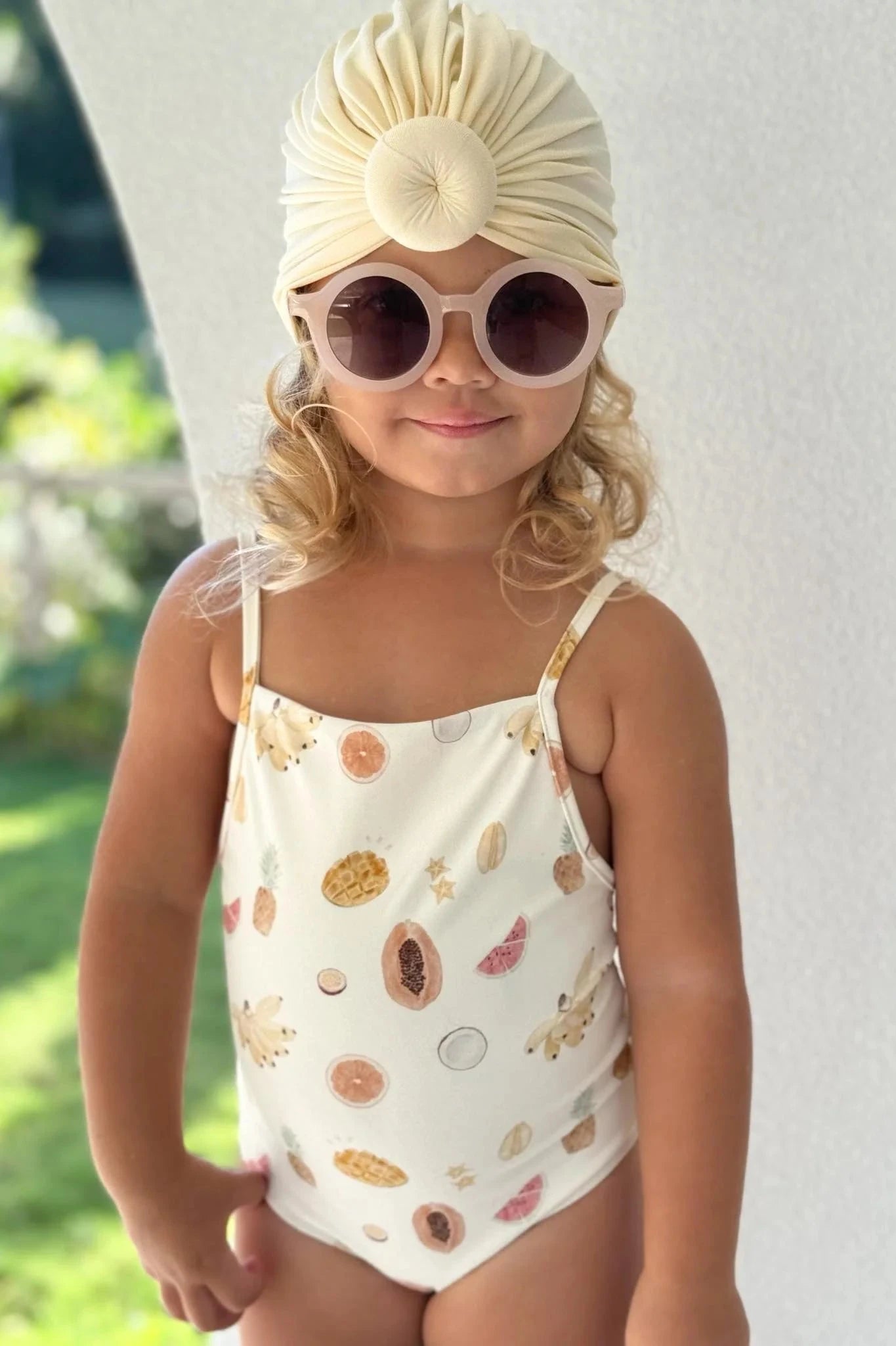 A young child wearing the EN. THE LABEL Tutti Frutti One Piece swimsuit with its fruit patterns and adjustable straps, a cream-colored turban, and large round sunglasses stands outdoors against a light-colored wall, smiling softly. The background includes a blurred view of greenery.