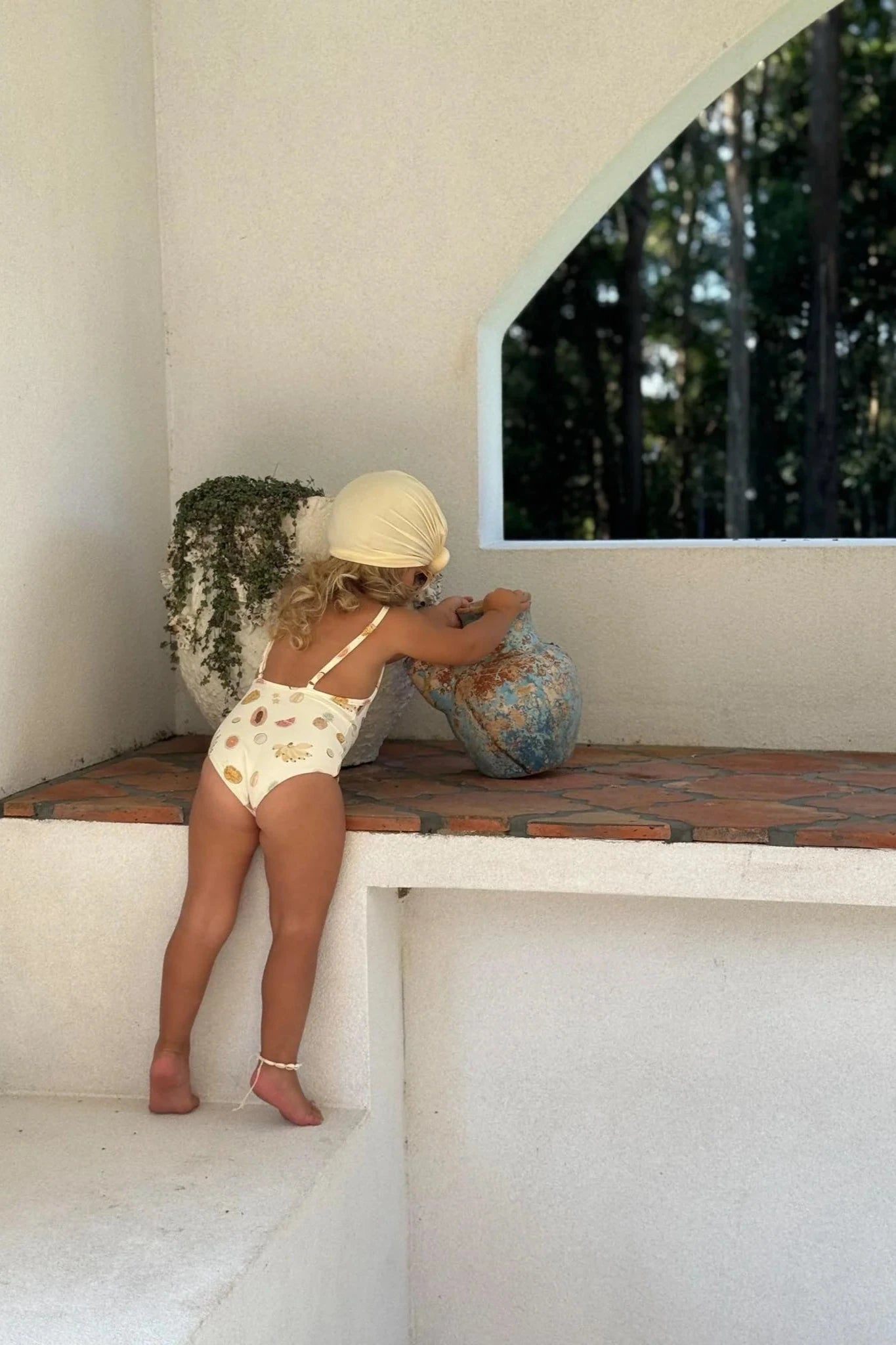 A young child with curly hair, wearing the Tutti Frutti One Piece chlorine-resistant toddler swimsuit from EN. THE LABEL and a headscarf, reaches up to touch a large, weathered stone planter on a raised ledge. Behind the child, through an arched window, forest scenery is visible. A potted plant sits nearby on the ledge.