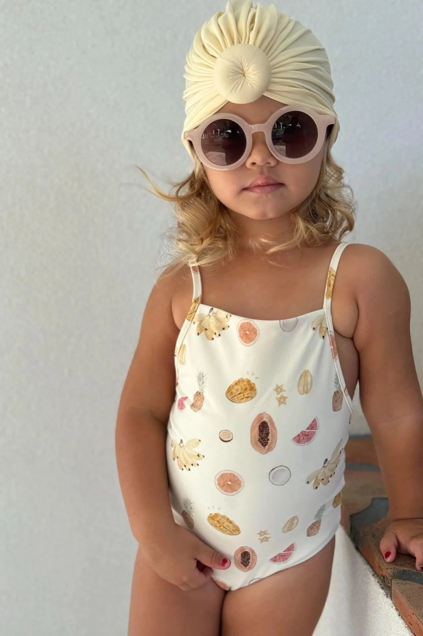 A young child with wavy blonde hair is wearing the Tutti Frutti One Piece, a cream-colored toddler swimsuit with fruit prints by EN. THE LABEL, paired with round sunglasses and a matching turban-style headband. This chlorine-resistant outfit features adjustable straps. They are posing against a plain white background.