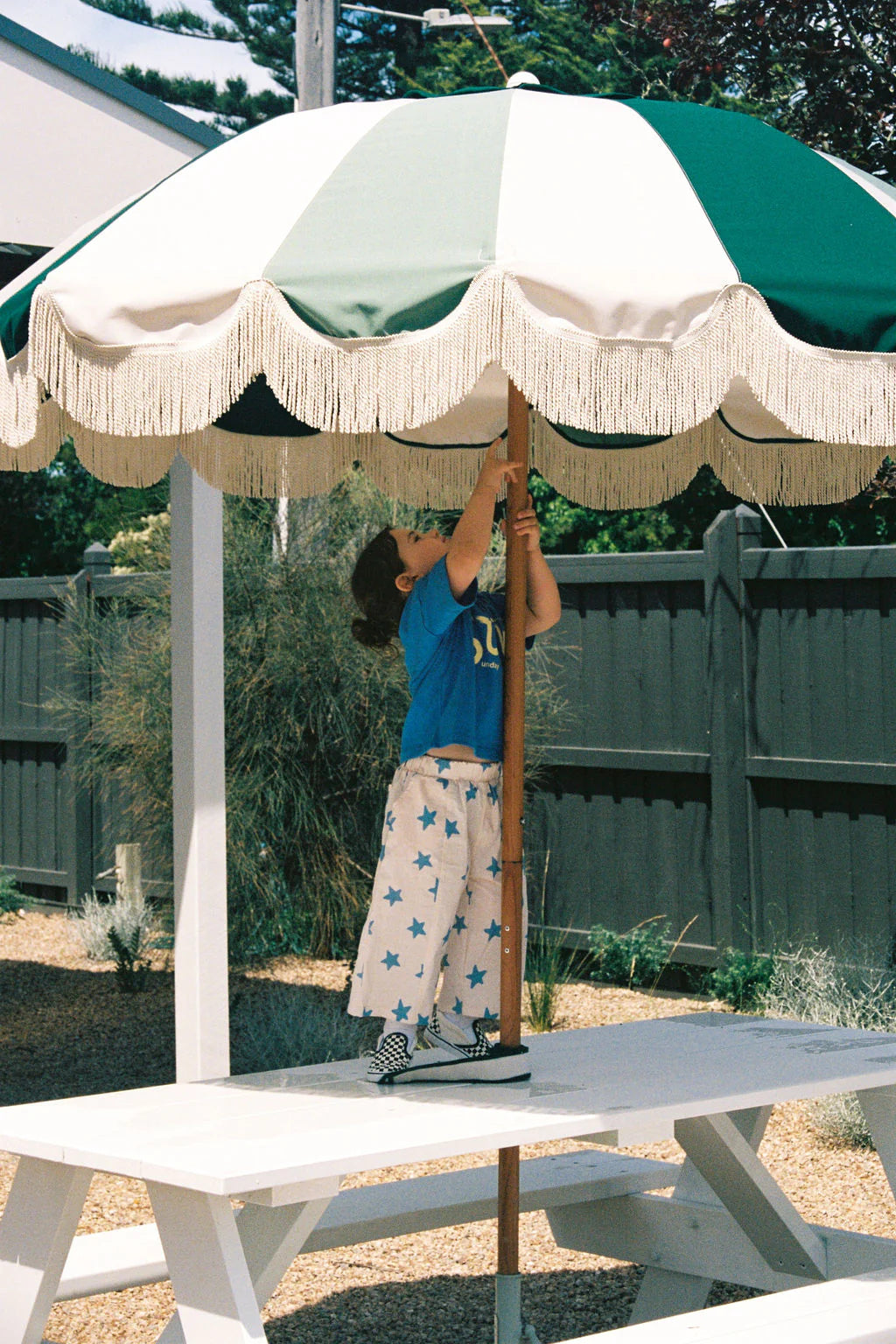 A child in oversized blue Il Sole Tee by SUNDAY SIBLINGS and star-patterned pants adjusts a large green and white fringed umbrella at an outdoor picnic area. A white bench and a black fence are visible in the background, illuminated by the sunny day.