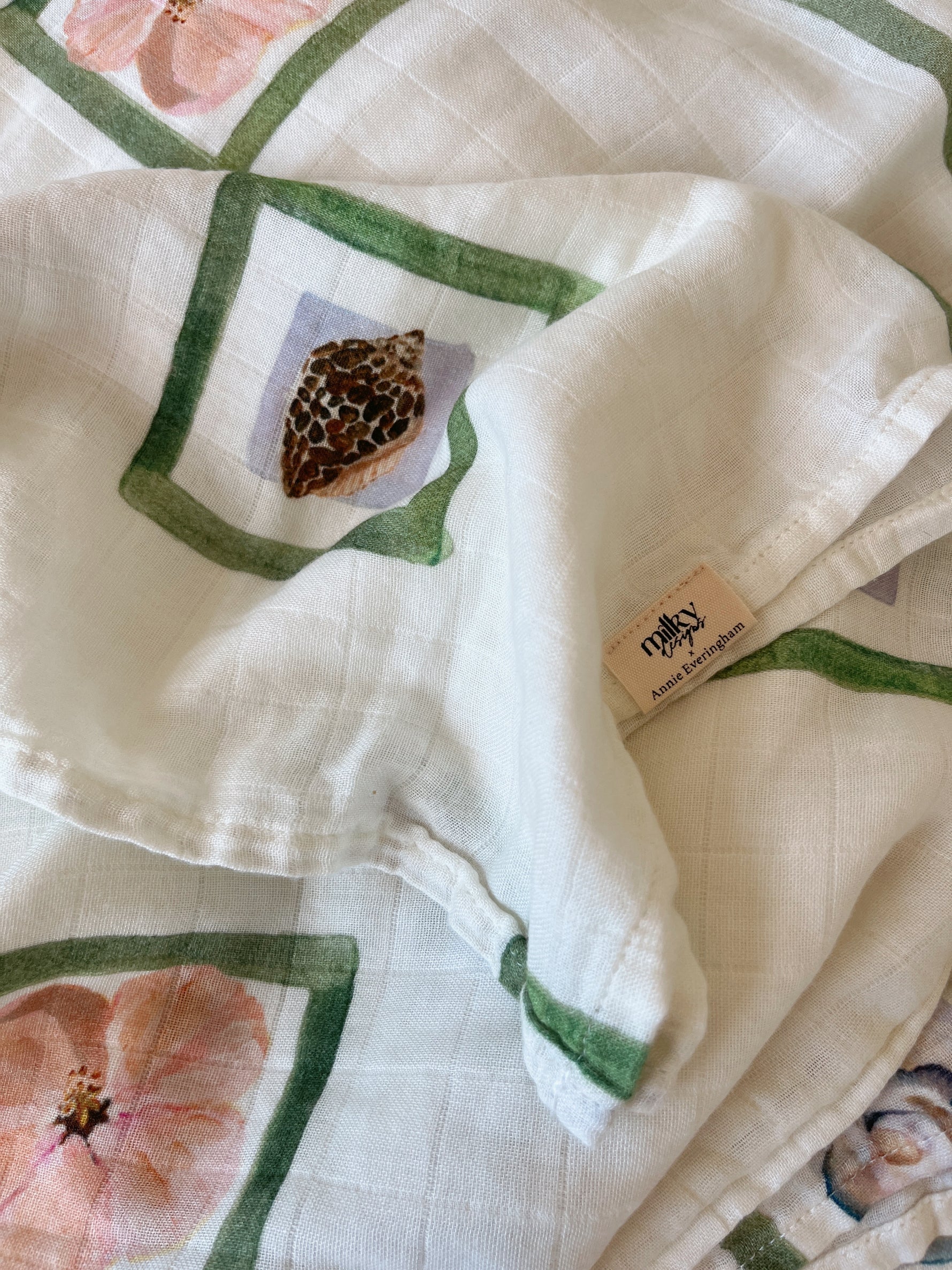 A detailed image of a white grid-patterned fabric features hand-painted designs with green frames, flowers, and a seashell. A small label reads "masey," followed by "Made in India," highlighting Milky Designs' Annie Wrap's attention to detail.