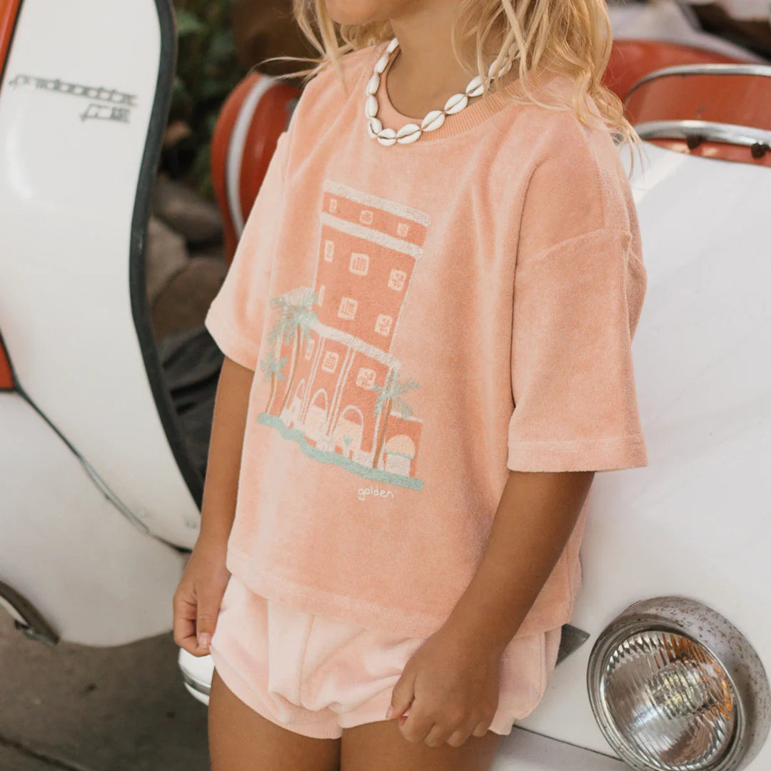 Near a classic white vintage car with a visible red interior, a child with long blond hair is wearing a peach-colored Room Service Mid Sleeve Tee Terry Towel Spanish Villa by GOLDEN CHILDREN, featuring a building illustration and adorned with a shell necklace. The scene evokes summer vibes reminiscent of afternoons at the Colony Hotel.