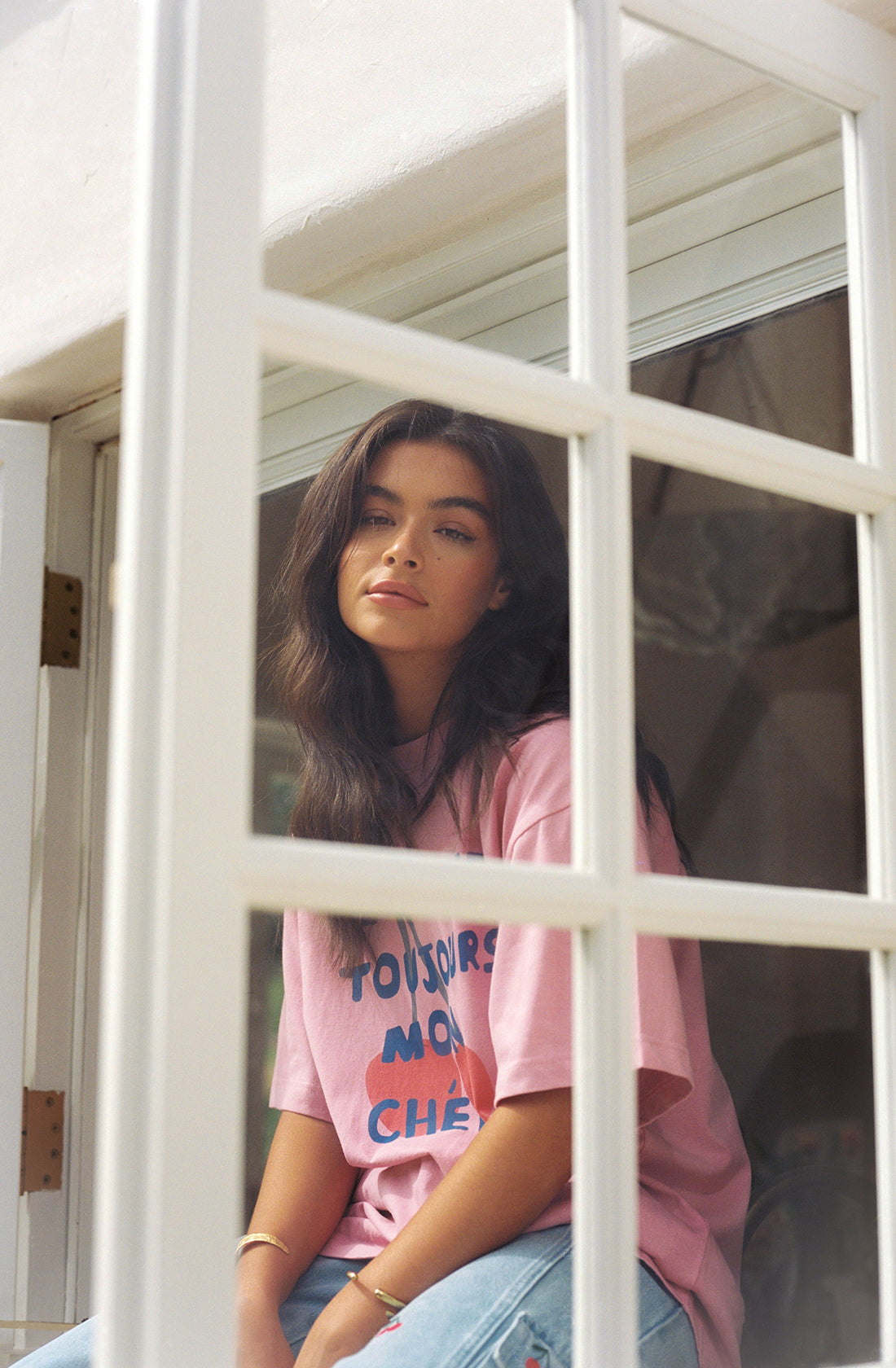 A young woman with long dark hair sits by a window, gazing calmly outside. She is wearing THE WHOLESOME STORE's ethically made Je T’aime Tee, a pink t-shirt with the words "TOUJOURS MON CHÉRI," and light blue jeans. The window's white wooden frame creates a grid pattern in the foreground.