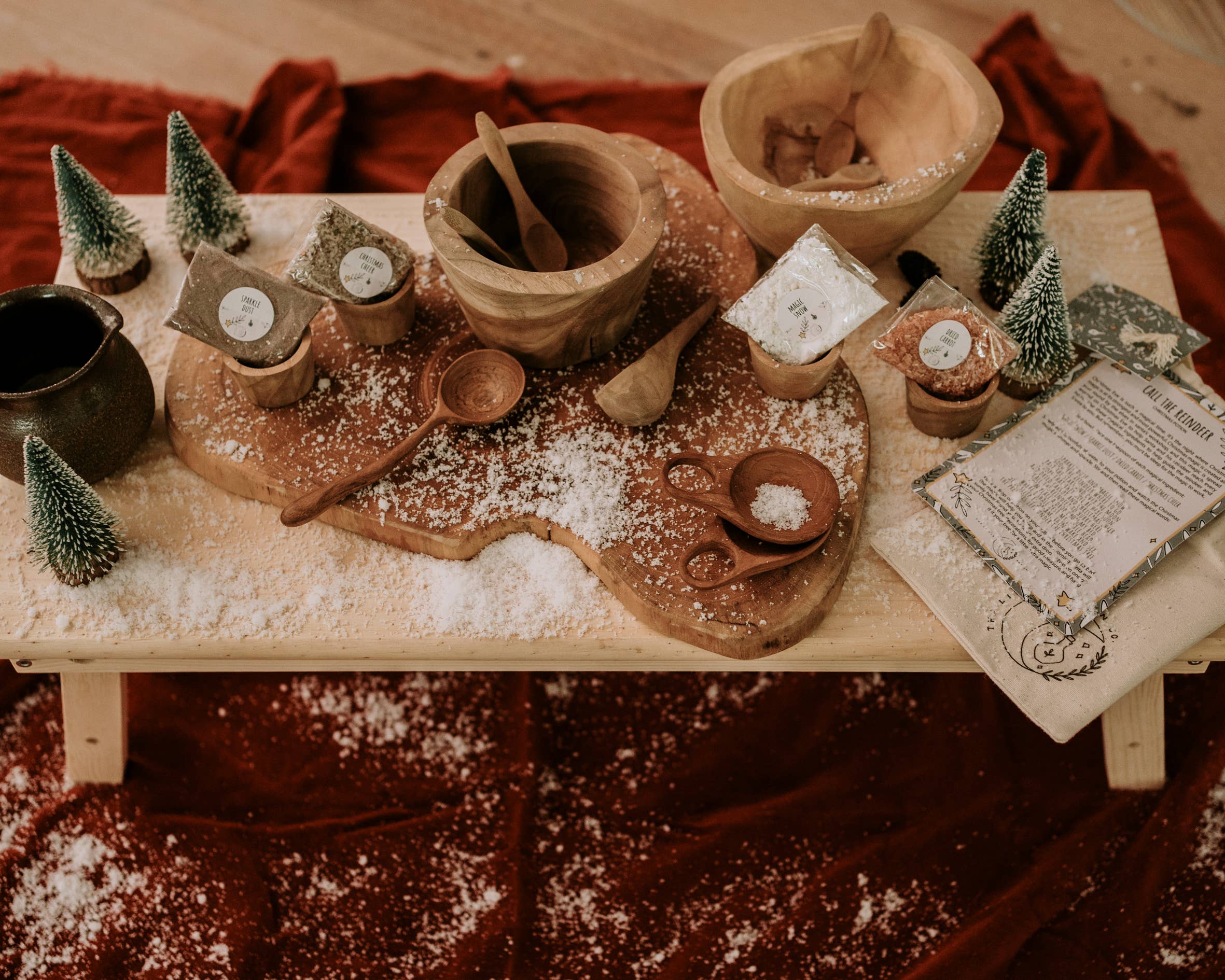 A cozy wooden table showcases the Xmas Eve Potion Pouch to Call the Reindeer by THE LITTLE POTION CO, surrounded by wooden bowls, spoons, and small packets nestled among tiny pine trees, dusted with flour or snow. A rich red cloth is draped underneath, creating a festive or rustic ambiance that brings to mind a Christmas potion kit.