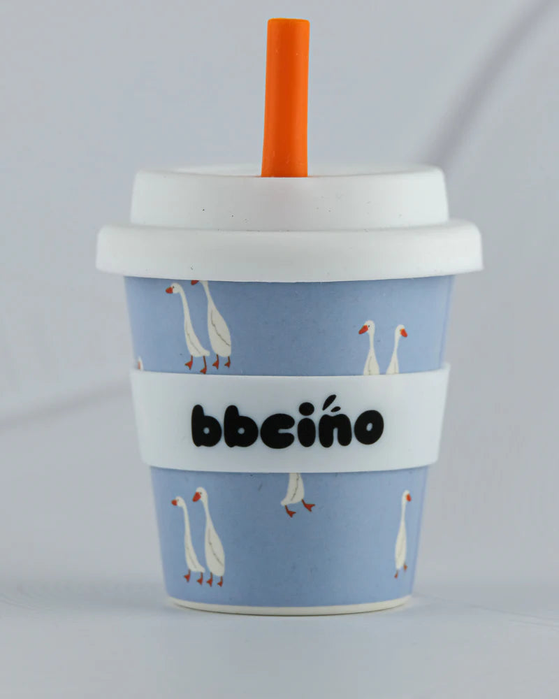 The Reusable Babycino Cup 120ml Loosey-Goosey by BBCINO is a blue silicone cup with a white lid and an orange straw. It features a charming pattern of white geese with orange beaks and includes a white band in the middle displaying "BBCINO" in black letters, making it ideal for eco-friendly babycino moments.