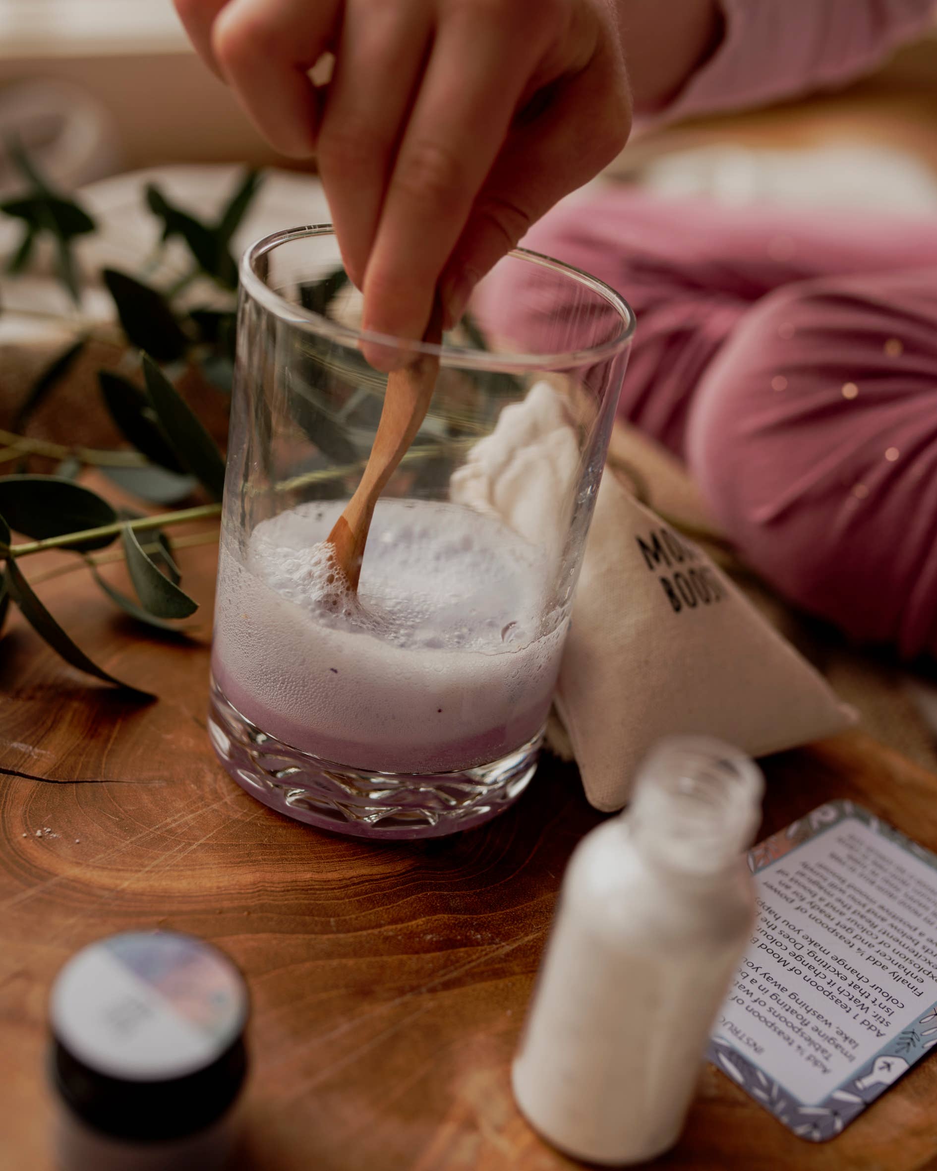 A person's hand stirs a frothy liquid in a glass with a small wooden spoon. Nearby are a cloth bag, a small jar, and a folded pamphlet, all on a wooden surface scattered with green leaves. The scene evokes the enchantment of THE LITTLE POTION CO's MINI Colour Mood Potion Kit (Colour Changing Kit), suggesting a whimsical and homemade skincare or wellness project.