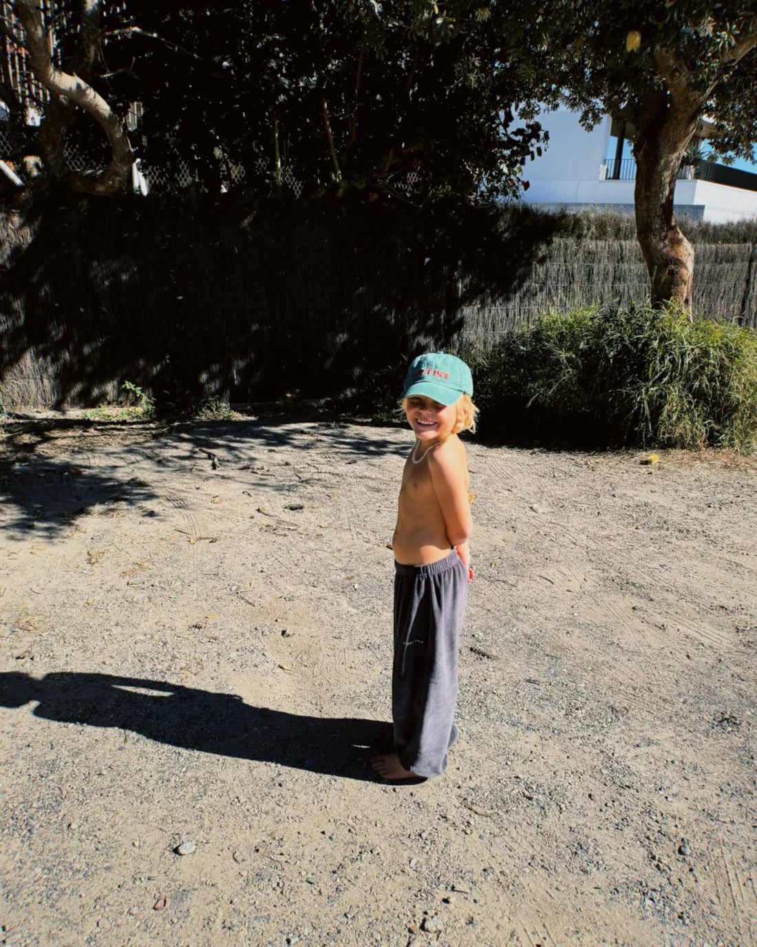 A child wearing the Ziggy Zaza ~ Artist Cap Forest Green by ZIGGY ZAZA and gray pants stands barefoot on a sunny, gravel-strewn path. Shirtless and smiling with hands behind their back, trees and a fence frame the scene while the cap's adjustable brass clasp subtly glints in the sun.