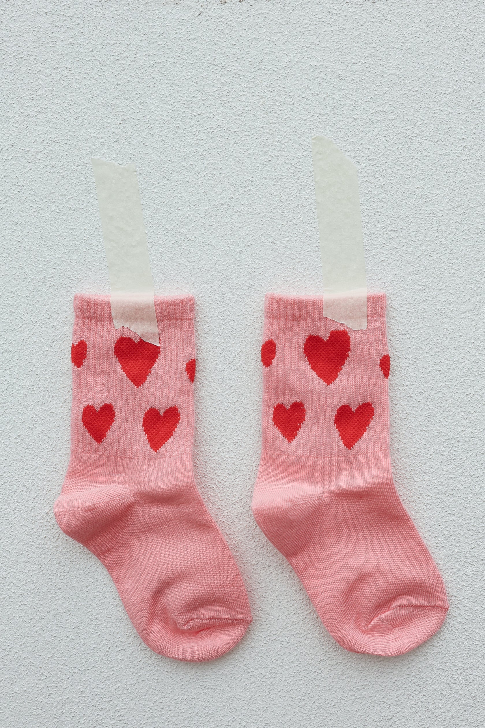 A pair of TINY LOVE CLUB's Burst Sock, kids' pink socks adorned with red heart patterns, is taped to a white wall. The hearts subtly vary in size and are evenly spaced across the top half of the socks, crafting a playful and cheerful design. These machine washable Burst Socks appear to be new and unworn.