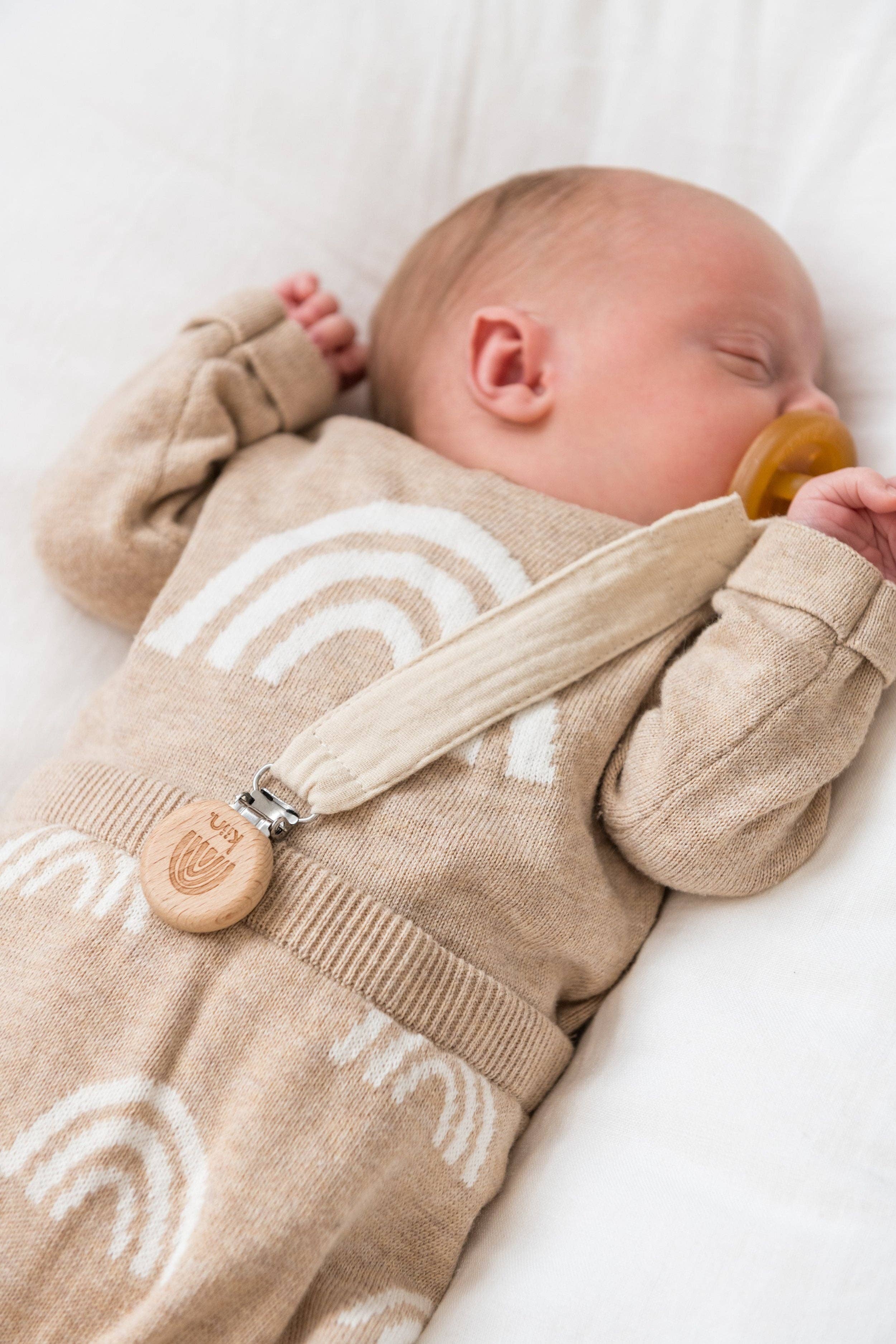 A baby in a beige outfit with white rainbows rests on a soft surface. Using the KIIN BABY Cotton Dummy Clip Oat, which features wooden details, this scene is ideal for a gender-neutral nursery and makes a perfect baby shower gift.