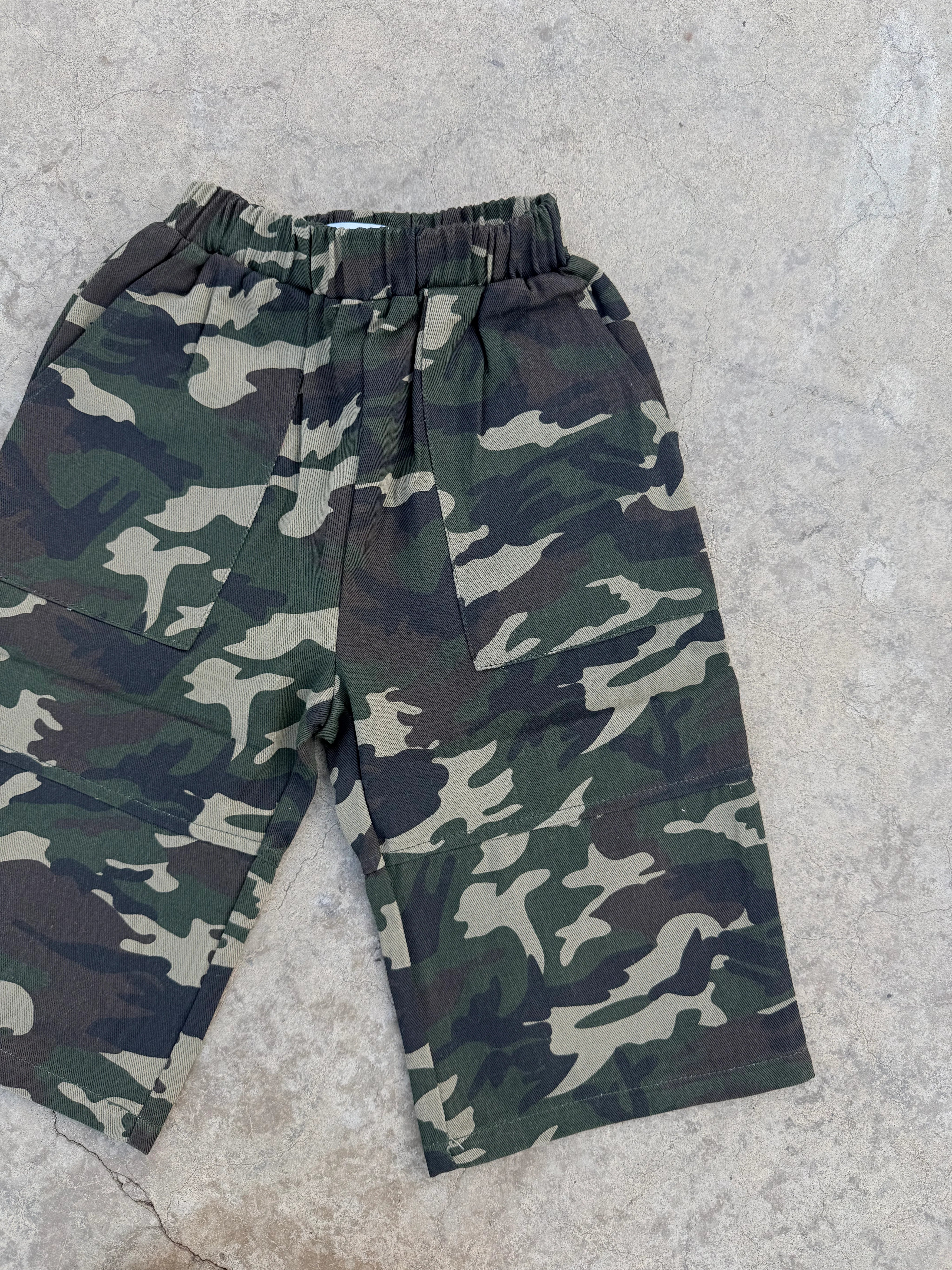 SAM THE LABEL’s “Sam the Label - The Cargo Pant Camo” kids’ cargos in green and brown camouflage, laid flat on a textured concrete surface.