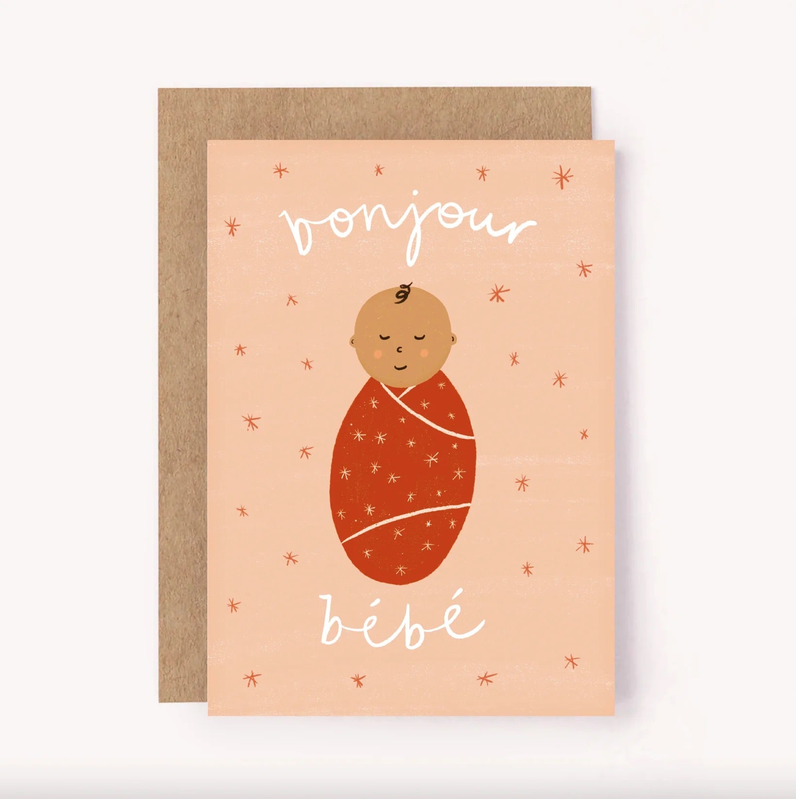 Introducing the Bonjour Bebe Pink Card - New Baby by LAUREN SISSONS STUDIO, a charming greeting card paired with a light brown envelope, perfectly suited for new parents. The card showcases an adorable illustration of a swaddled baby in a red blanket with closed eyes. Set against a peach-colored background adorned with small star patterns, the text "bonjour bébé" is elegantly scripted in white cursive at both the top and bottom.