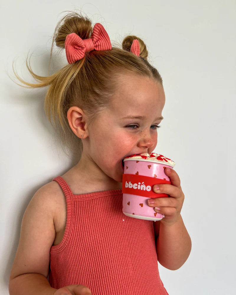 A young child with two hair buns and red hair bows joyfully licks whipped cream from a BBCINO Reusable BambinoCino Cup 240ml Sweetheart, designed with heart patterns, while wearing a sleeveless pink top against a plain white background—an eco-friendly alternative to single-use cups.