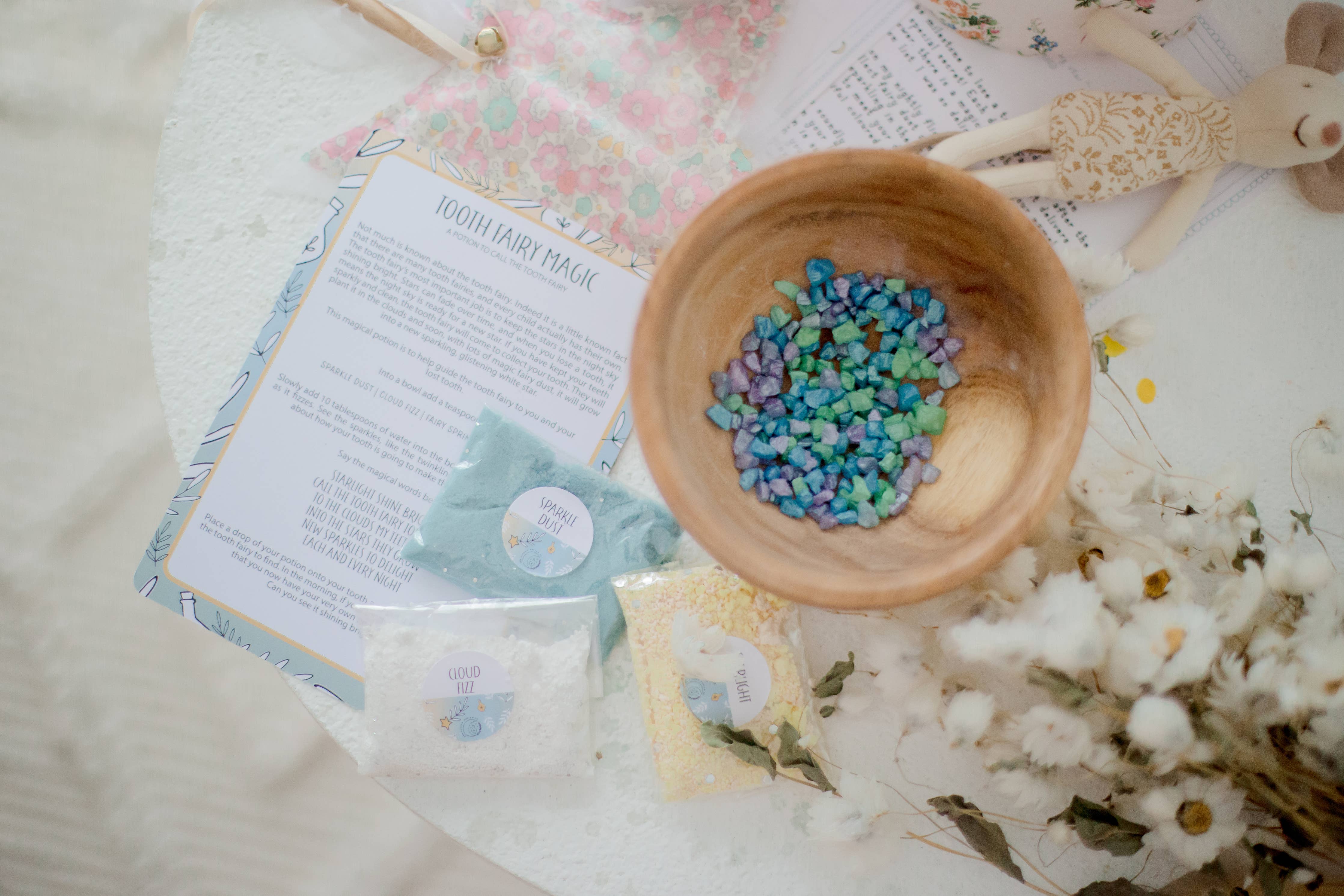 A table features a wooden bowl filled with colorful stones, accompanied by craft supplies such as bags labeled "Unicorn Dust" and "Gold Star," alongside a paper titled "1001 Fairy Magic." A floral notebook, cloth rabbit toy, and cotton flowers are also present, suggesting the creation of the enchanting Tooth Fairy Magic Potion Pouch by THE LITTLE POTION CO.