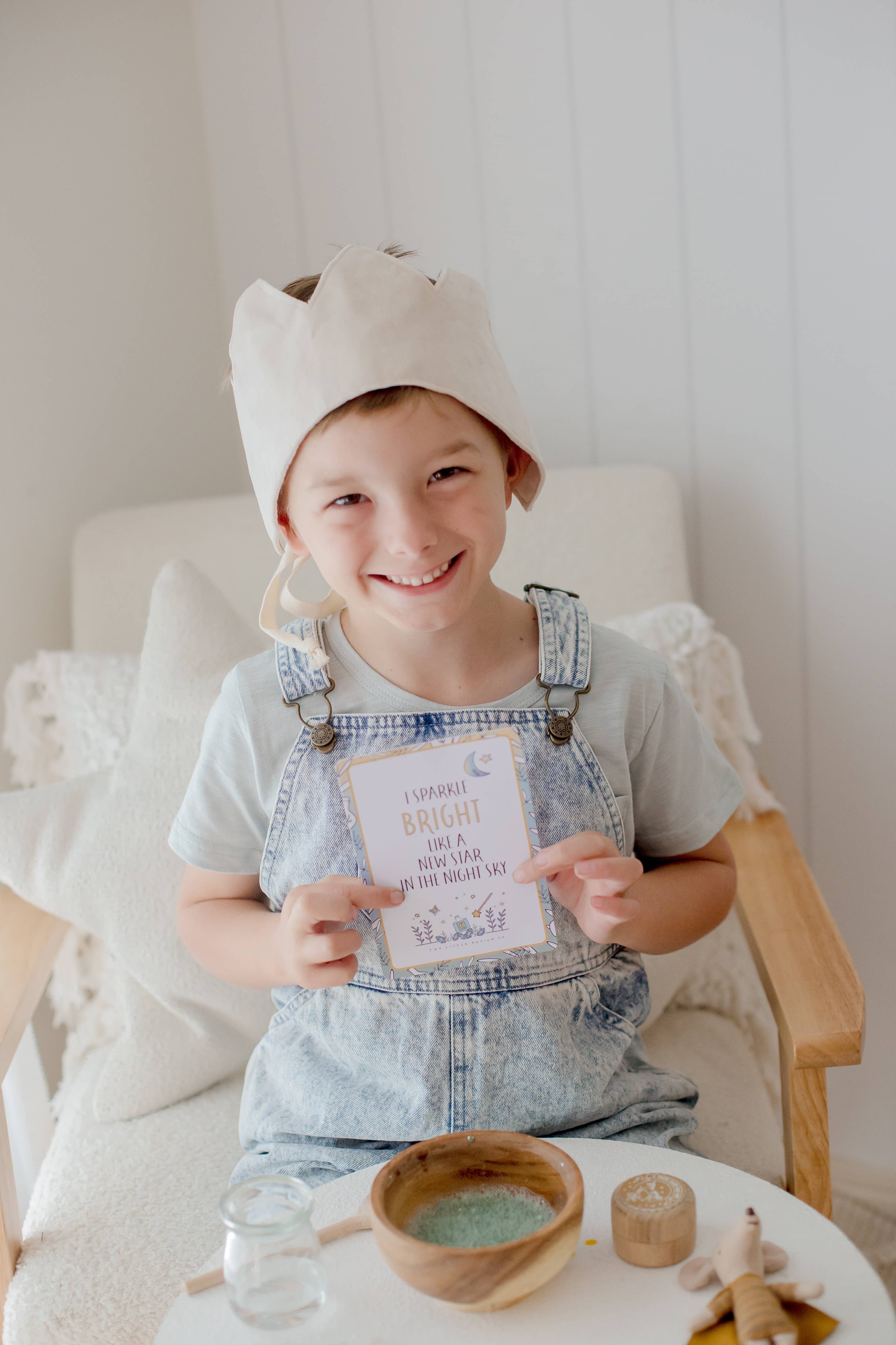 A joyful child wearing a paper crown sits in a chair, holding a card that reads "Sparkle bright and be fun, you are the bright one." The child, dressed in denim overalls, is surrounded by craft supplies and a Tooth Fairy Magic Potion Pouch filled with sparkly ingredients from THE LITTLE POTION CO.