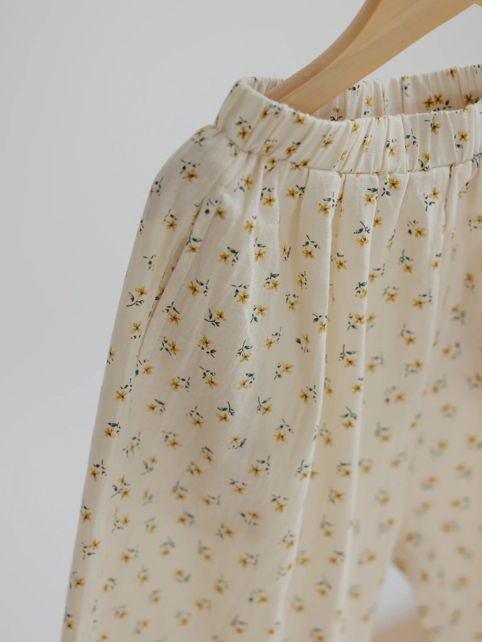 A close-up image of the VALENCIA BYRON BAY Rio Pant Marigold, featuring a small yellow floral pattern in a delightful beige hue. These durable pants are made from 100% cotton and feature an elastic waistband, hanging on a light wood hanger against a white background.