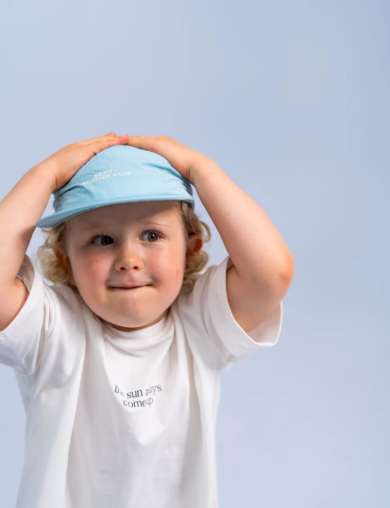 A young child with curly hair stands against a light blue background, wearing the Summer Club Cap : Ocean from SONNY LABEL, along with a white T-shirt that reads "The sun always comes up." The mini explorer playfully rests their hands on their head, looking slightly upward.