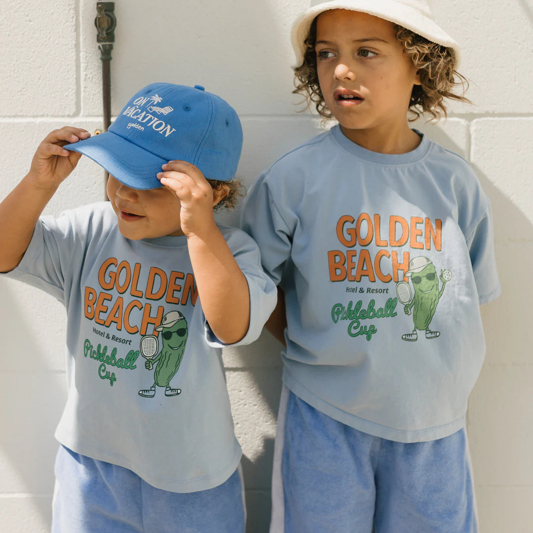 Two children stand side by side in relaxed fit "Golden Beach Mid Sleeve Tee Palm Blue" from GOLDEN CHILDREN, paired with matching blue outfits. One adjusts a blue cap while both smile naturally against the simple white wall, exuding casual charm.