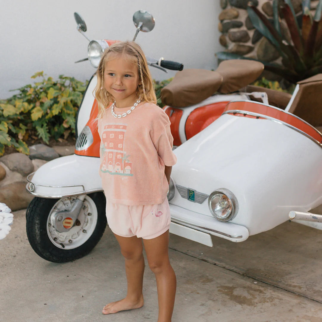 A young girl with blonde hair stands smiling in front of a vintage orange and white scooter with a sidecar. She is wearing a Room Service Mid Sleeve Tee Terry Towel from GOLDEN CHILDREN in the color Spanish Villa and matching shorts, with lush plants flourishing in the background.