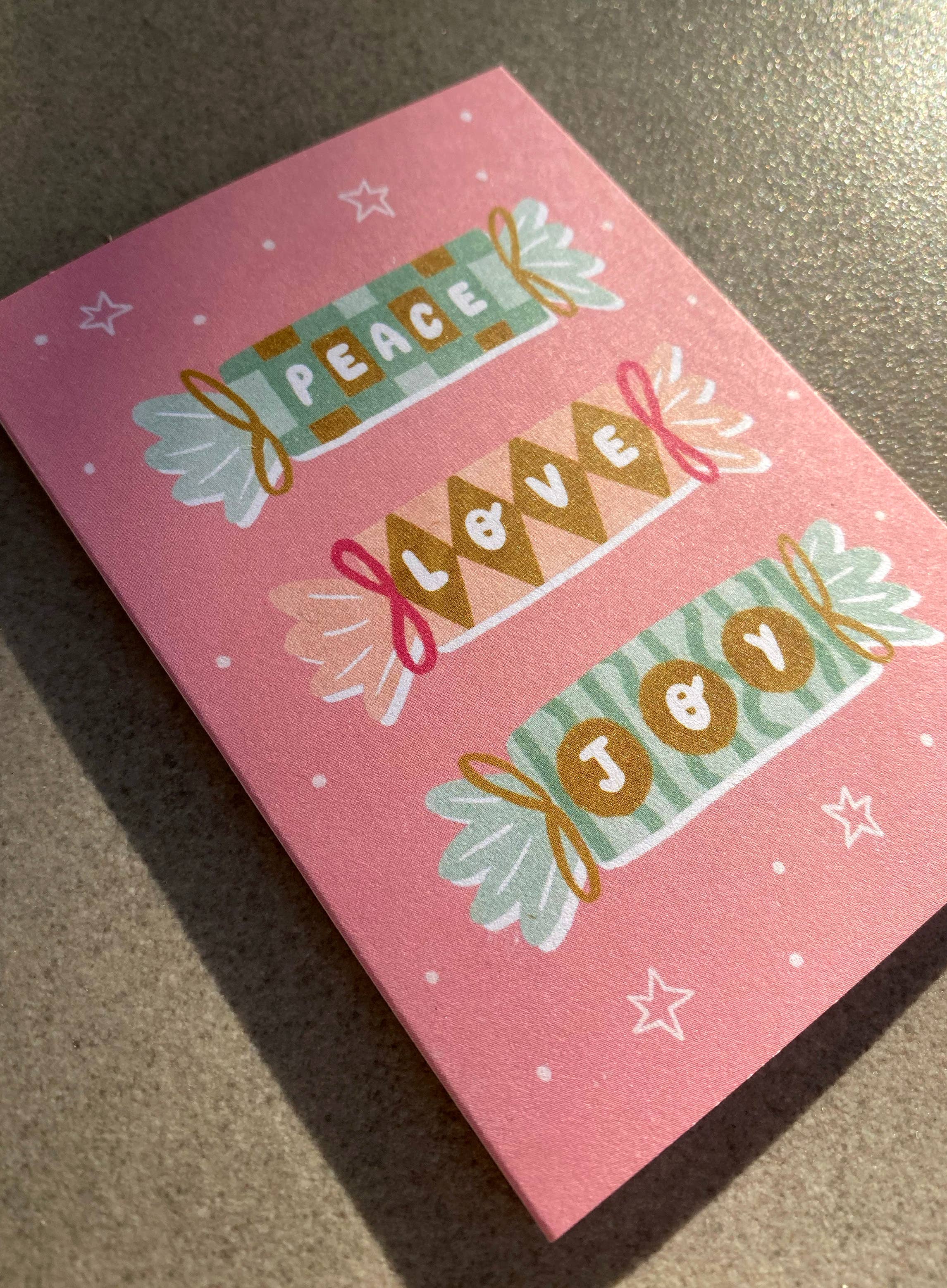 The Peace Love Joy Christmas Crackers Mini Card by LAUREN SISSONS STUDIO features a pink design with decorative text in the shapes of three colorful candies, spelling out "Peace," "Love," and "Joy." Made from recycled paper, it is embellished with small white dots and stars, resting on a flat surface where sunlight casts a soft shadow.