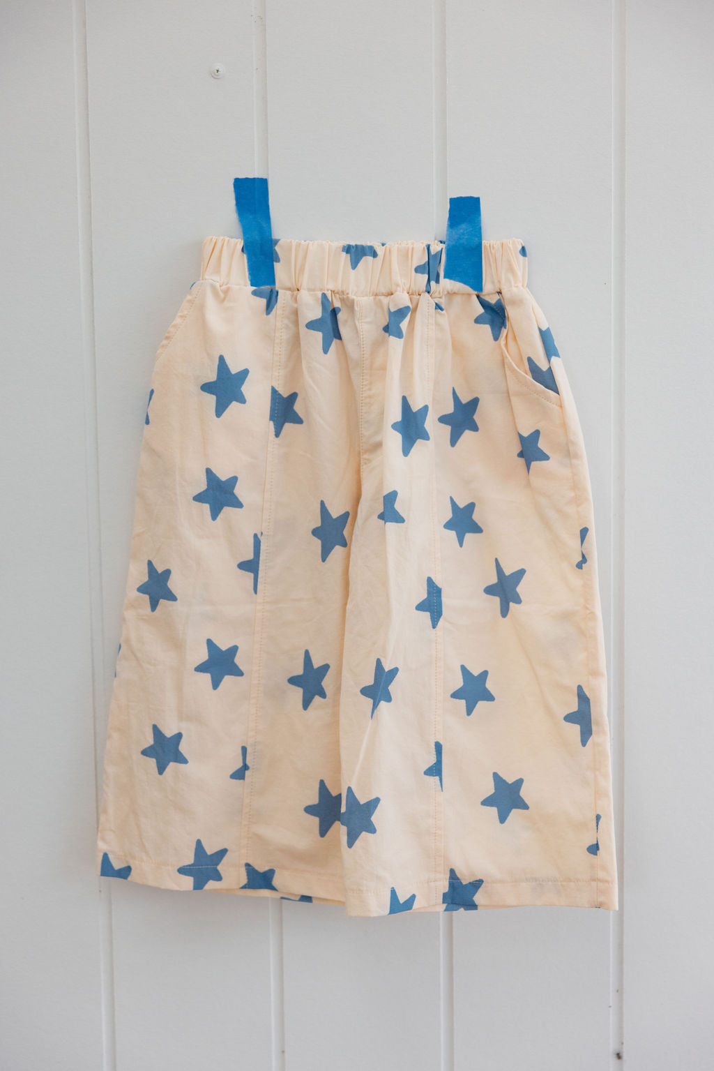 The SUNDAY SIBLINGS - Dusty Star Pants, featuring a beautiful beige and blue star pattern, are taped to a white wall with blue tape at the waistband. Made of cotton with a gathered, elastic waistband, they exude a relaxed and simple charm.