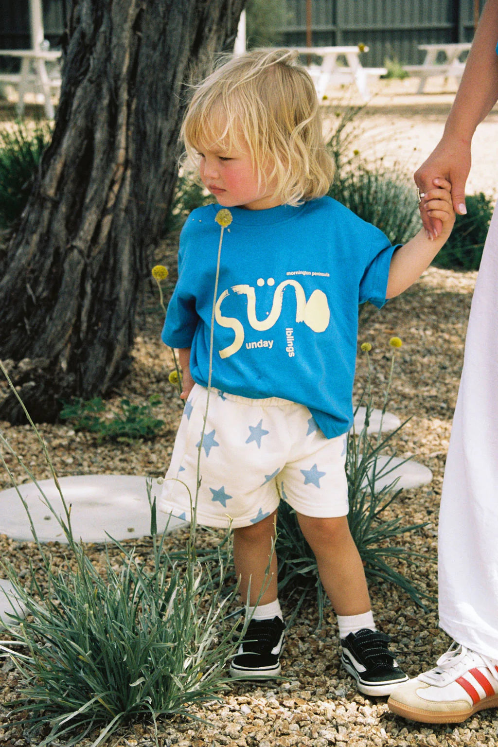 A young blond child stands outdoors holding an adult's hand, wearing a bright blue shirt with yellow text and SUNDAY SIBLINGS Kiddo Shorts in blue, made of soft French terry cotton with an elastic waistband. Nearby, there's a tree surrounded by gravel and grass. The child's shoes are black and white.